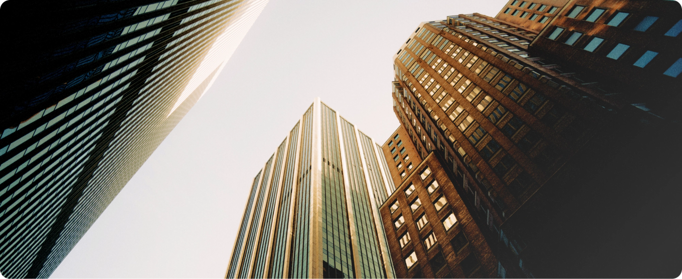 Upward view of a cityscape showcasing tall modern skyscrapers against a grey sky. The scene feels dynamic and urban.