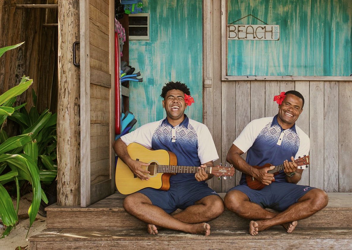 Two smiling musicians playing guitar and ukulele outside a rustic beachfront hut, surrounded by tropical greenery and palm trees.