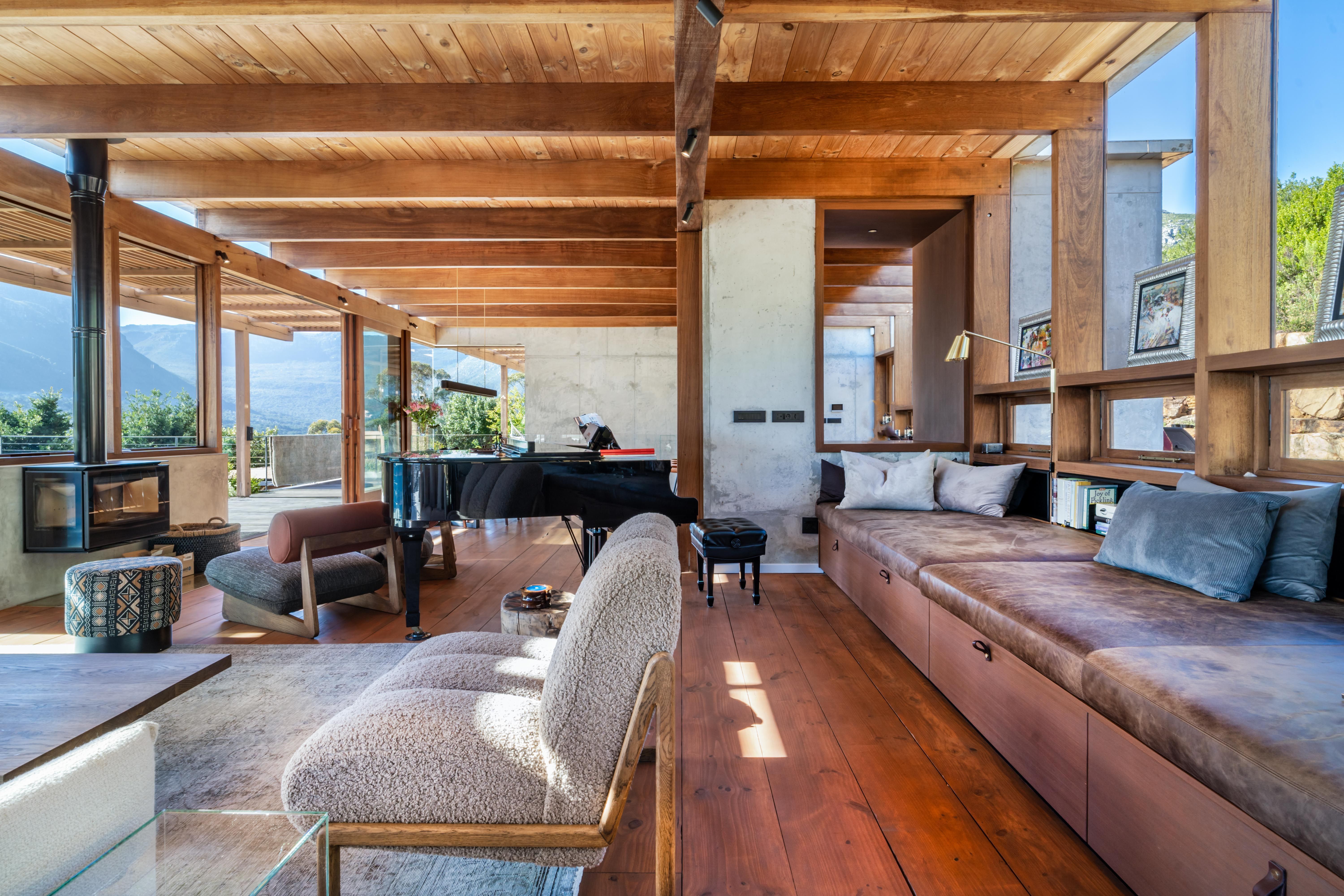 Living room interior showing the materials in conversation: concrete, dark wood beams, stone fireplace, mountain through floor-to-ceiling glass