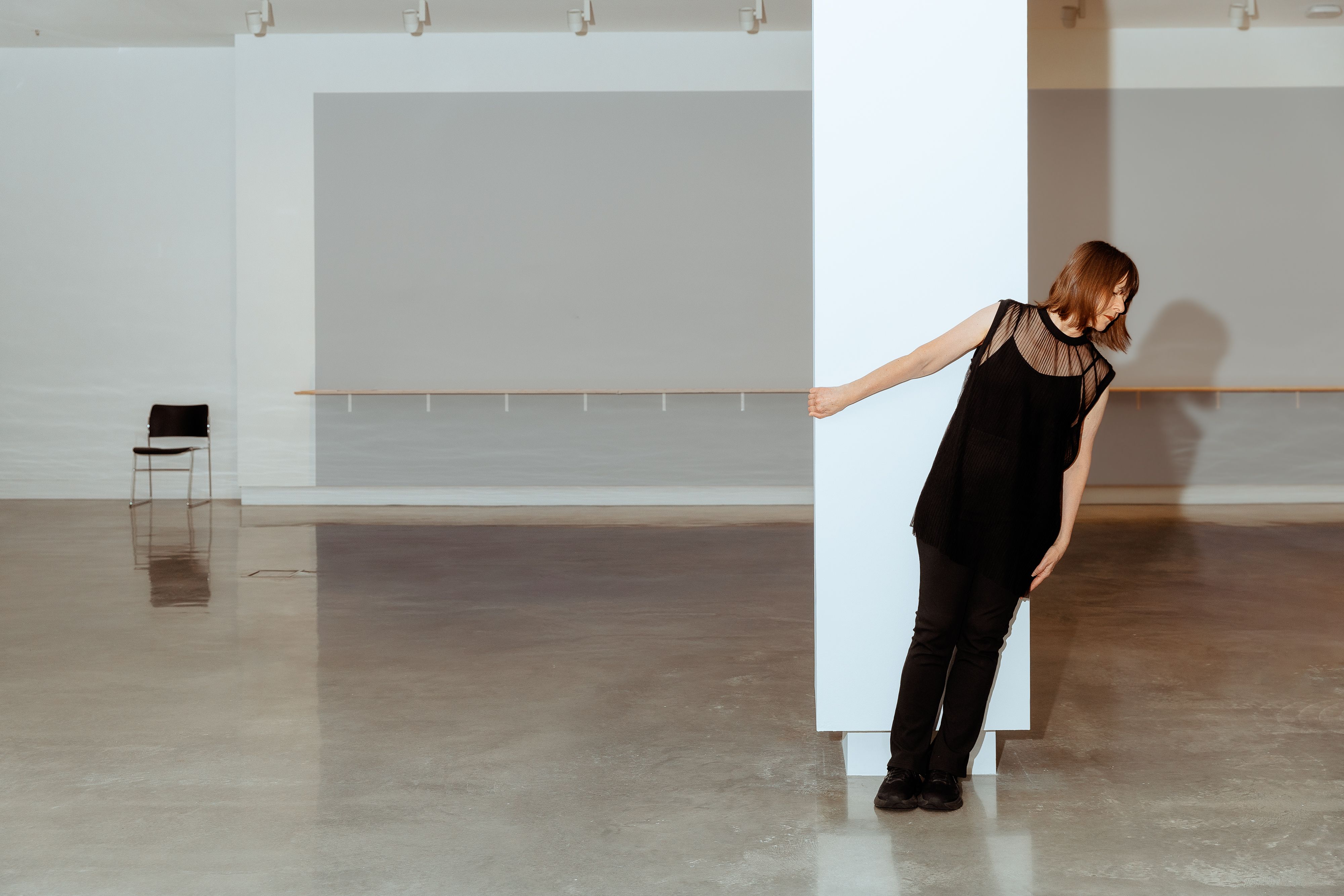 A woman in all black leans against a white column in an empty gallery space.