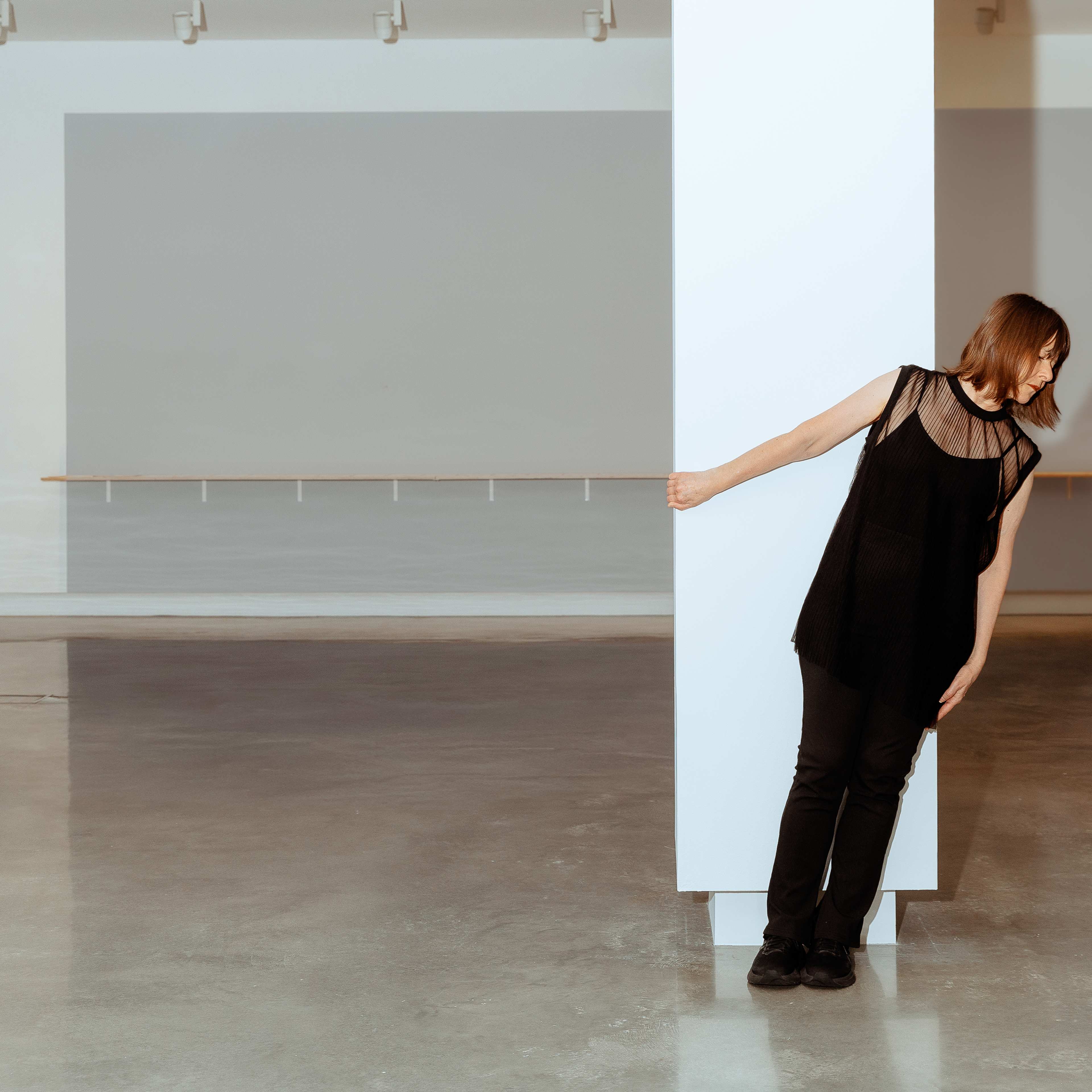 A woman in all black leans against a white column in an empty gallery space.