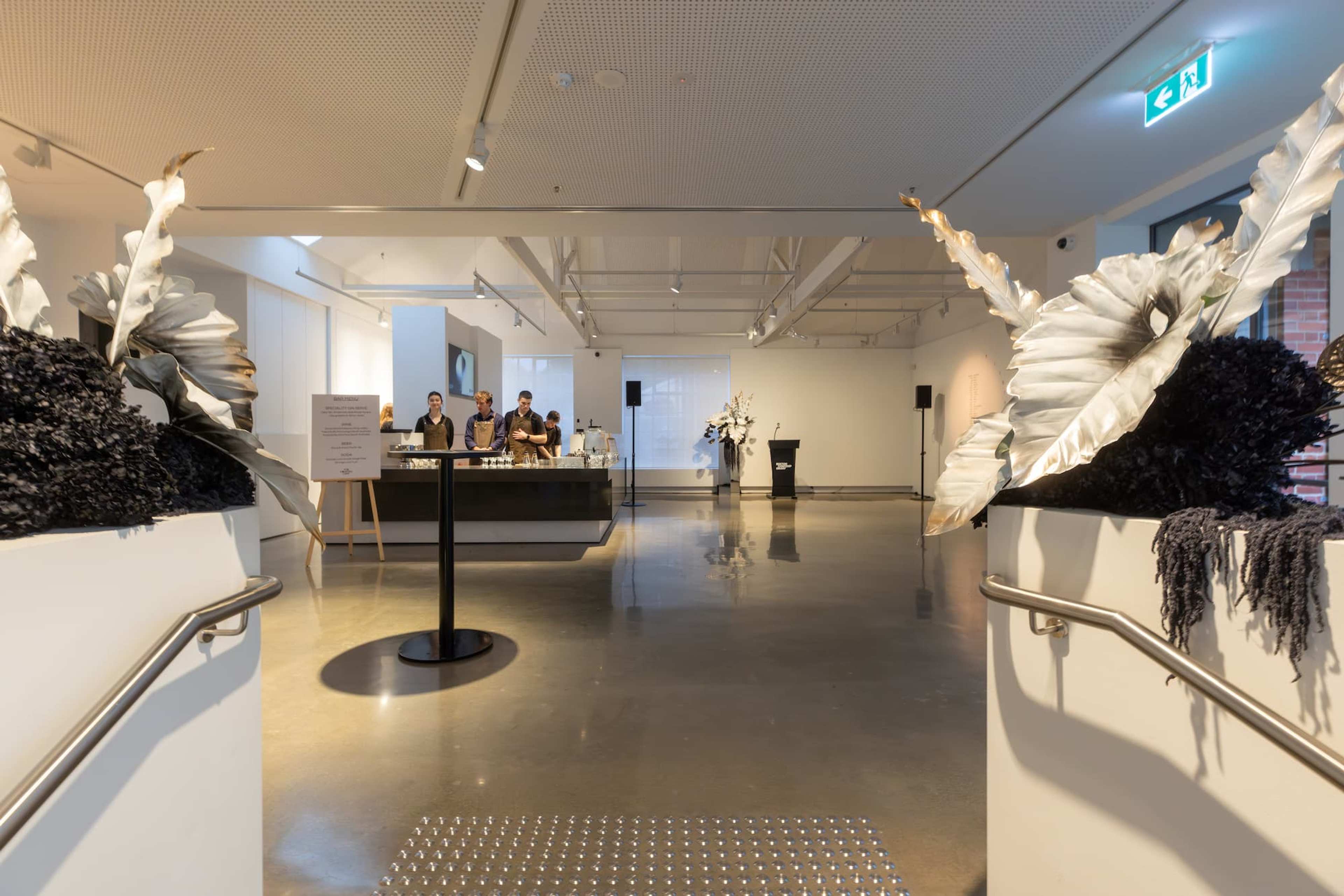 A modern art gallery lobby with shiny floors, white walls, large decorative plants, and a reception desk where several people are standing. Contemporary art pieces are displayed in the background.