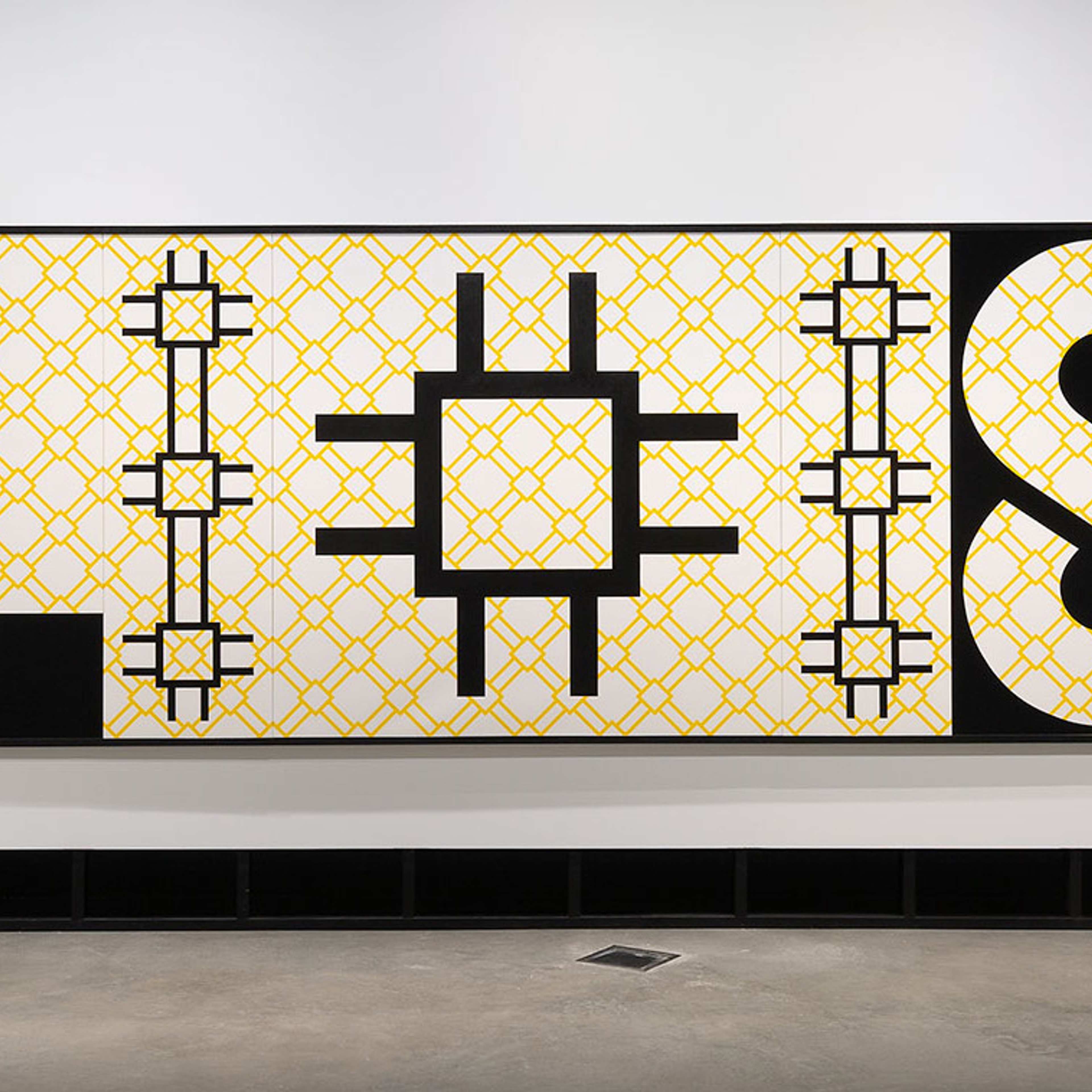 A gallery wall displays twelve framed geometric yellow artworks arranged in front of a net-like installation. The floor is polished, and a simple wooden bench sits in the foreground.