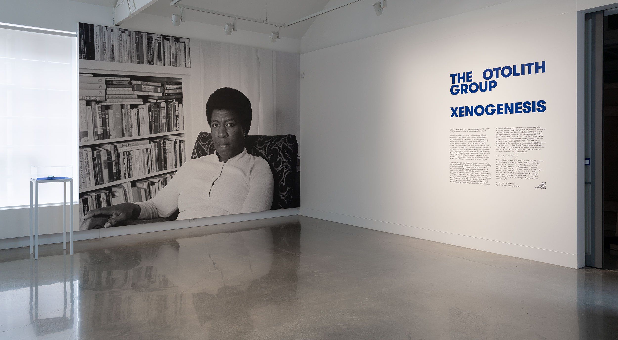 A gallery space features a large black-and-white portrait of a woman seated in front of bookshelves. On an adjacent white wall, blue text reads The Otolith Group Xenogenesis with an exhibition description below.