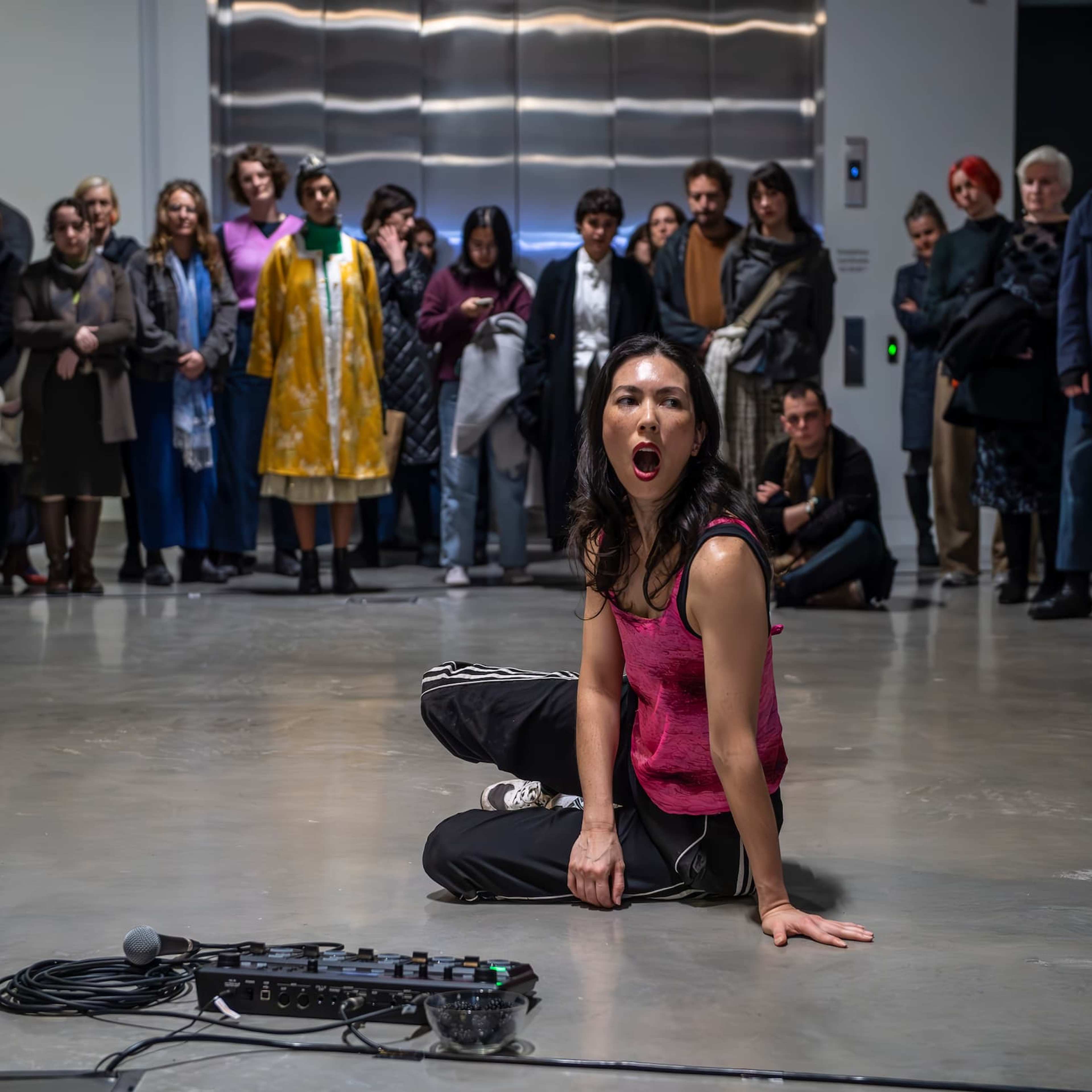A performer in a pink tank top sits on the floor, mouth open, in front of a crowd watching attentively in a gallery space. A microphone and audio equipment are on the floor nearby.