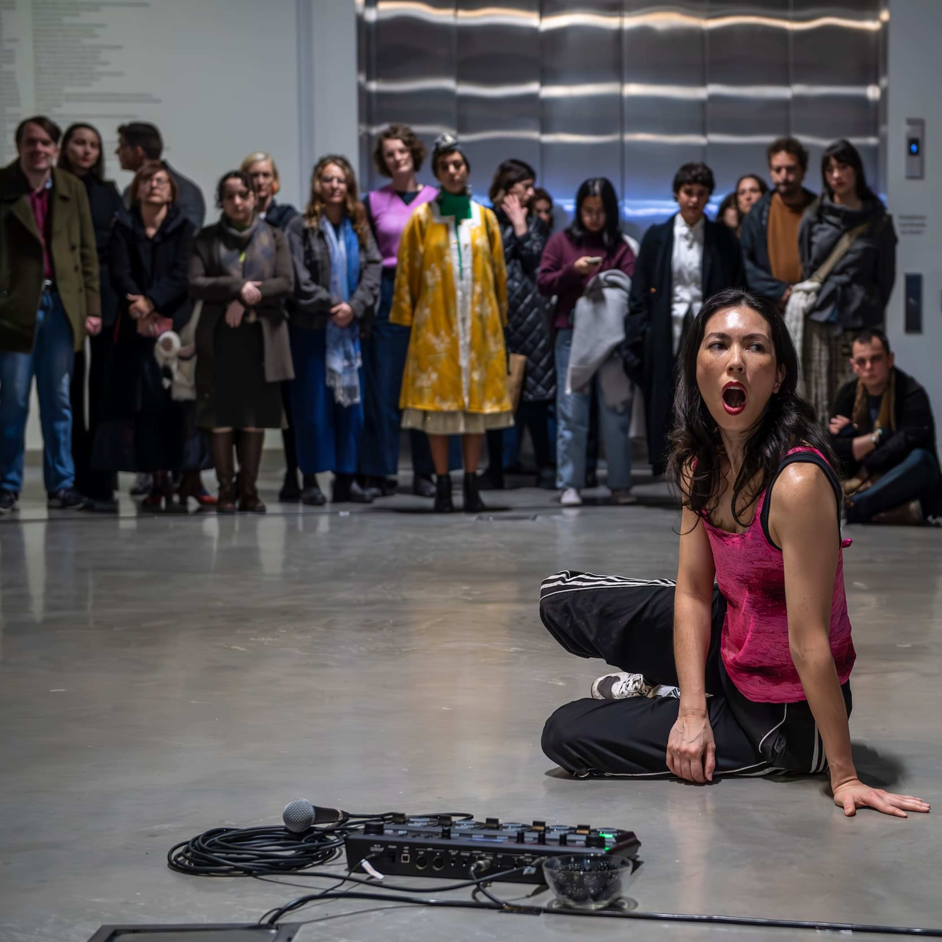 A performer in a pink tank top sits on the floor, mouth open, in front of a crowd watching attentively in a gallery space. A microphone and audio equipment are on the floor nearby.