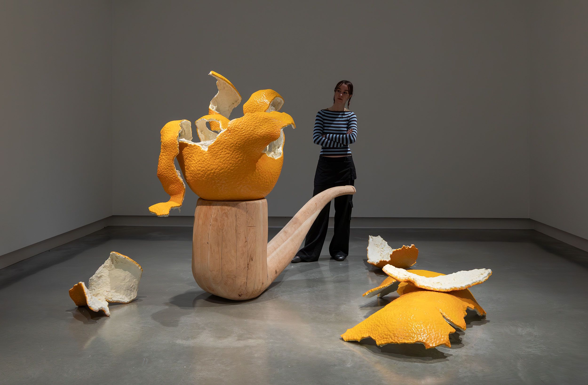 A person stands in a gallery, observing a large sculpture of a wooden pipe with oversized, broken orange peel pieces emerging from and scattered around it on the floor.