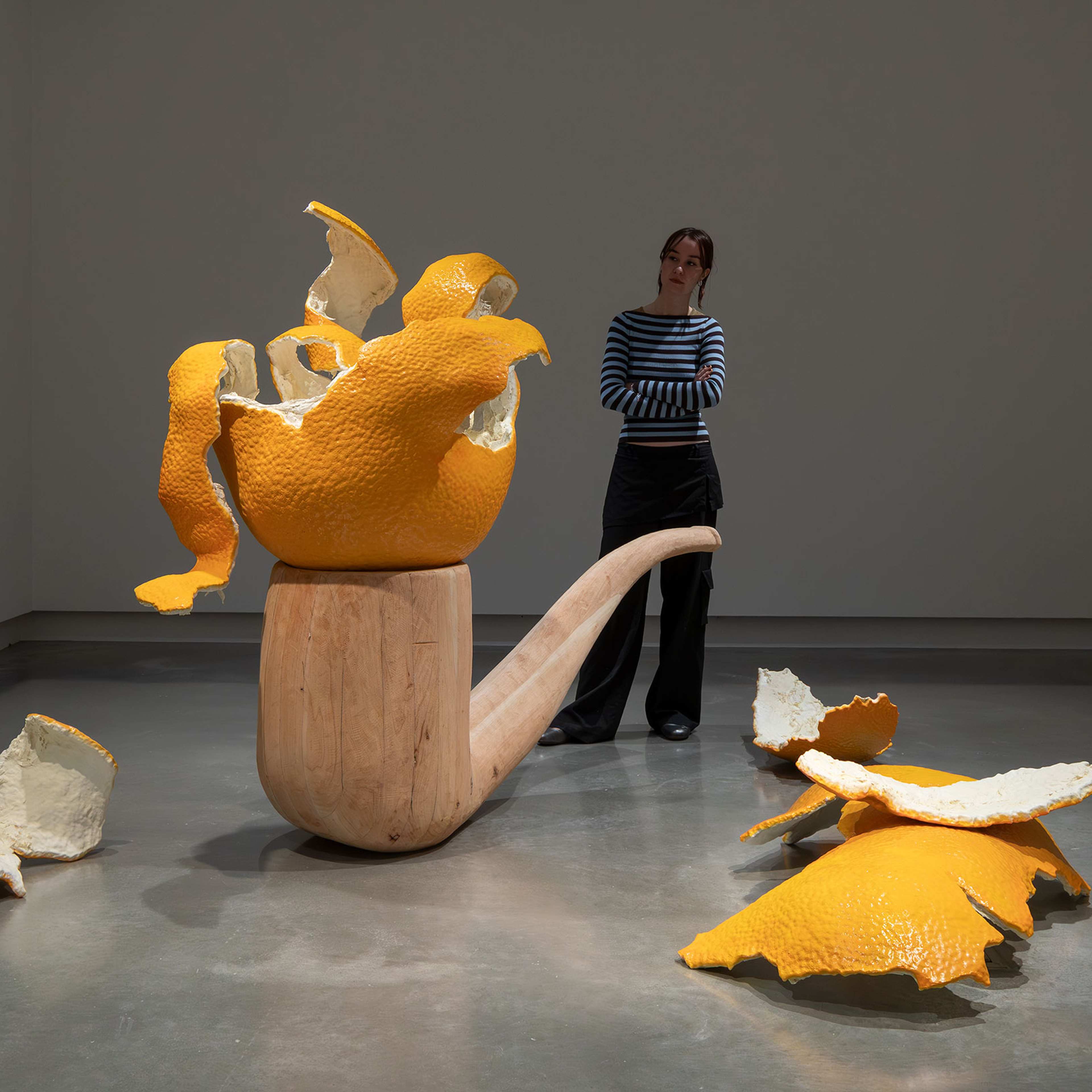 A person stands in a gallery, observing a large sculpture of a wooden pipe with oversized, broken orange peel pieces emerging from and scattered around it on the floor.