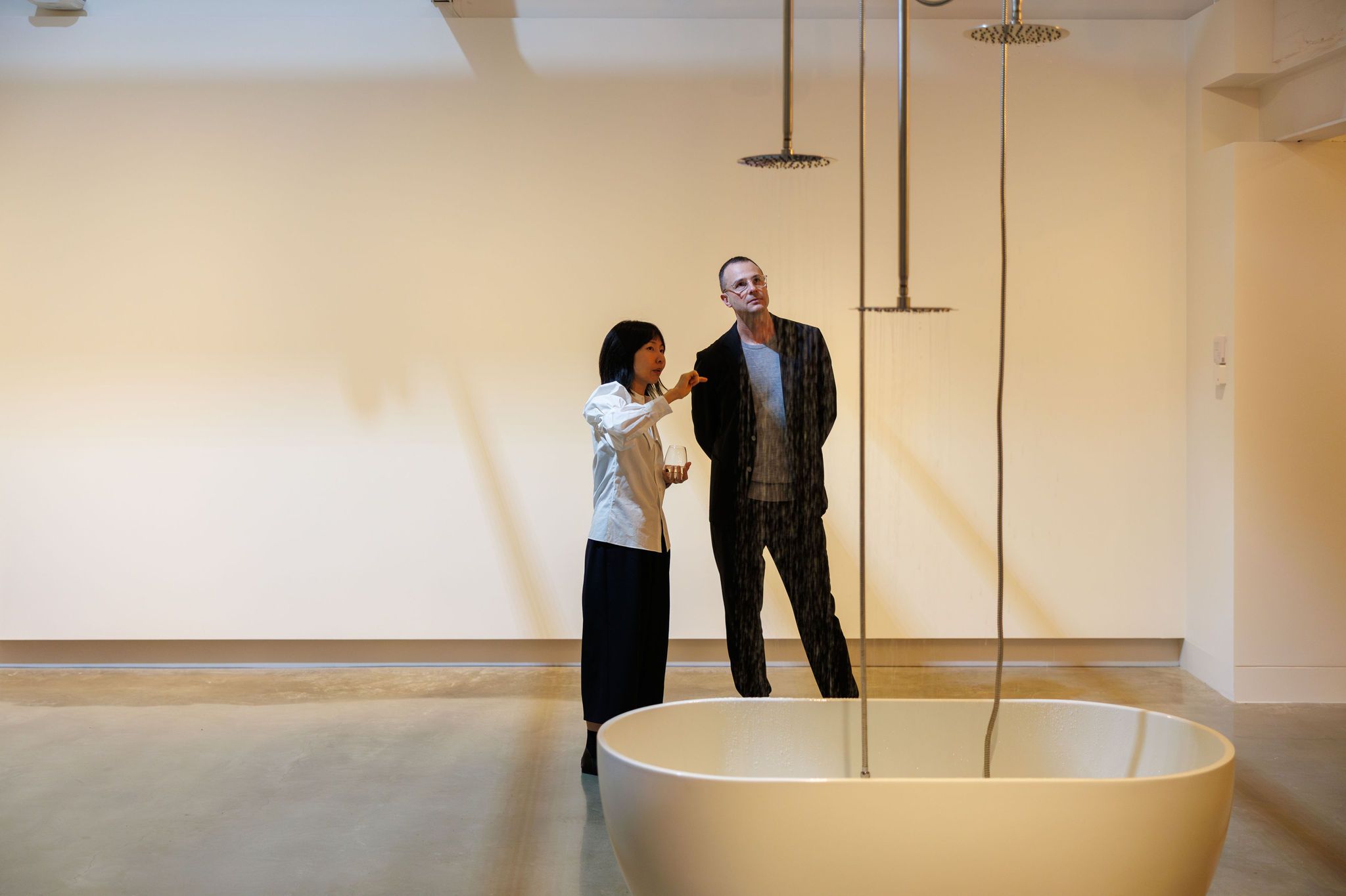 A woman gestures while talking to a man in an art gallery, both observing water falling from ceiling-mounted shower heads into a large white basin. The background is minimal and softly lit.