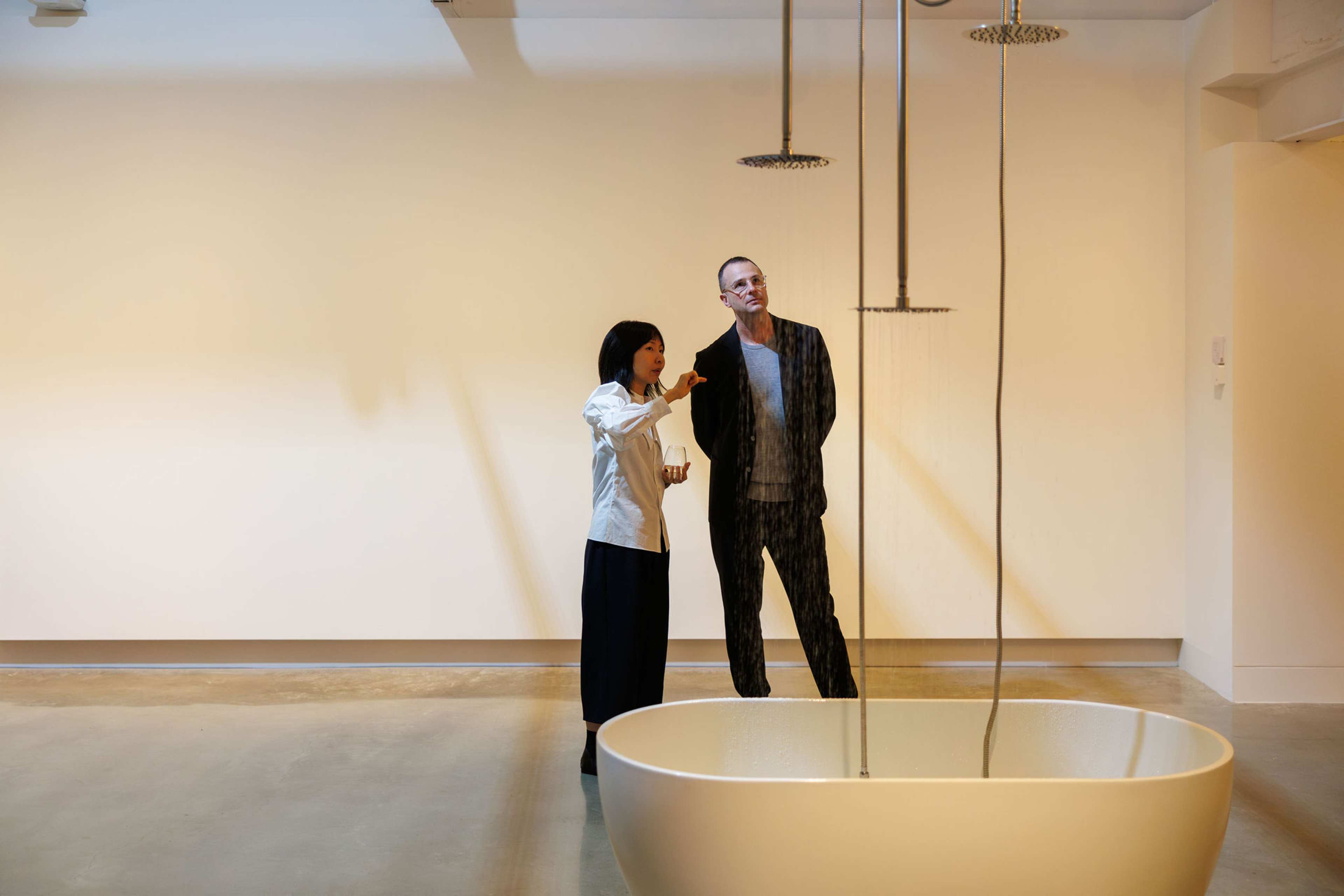 A woman gestures while talking to a man in an art gallery, both observing water falling from ceiling-mounted shower heads into a large white basin. The background is minimal and softly lit.