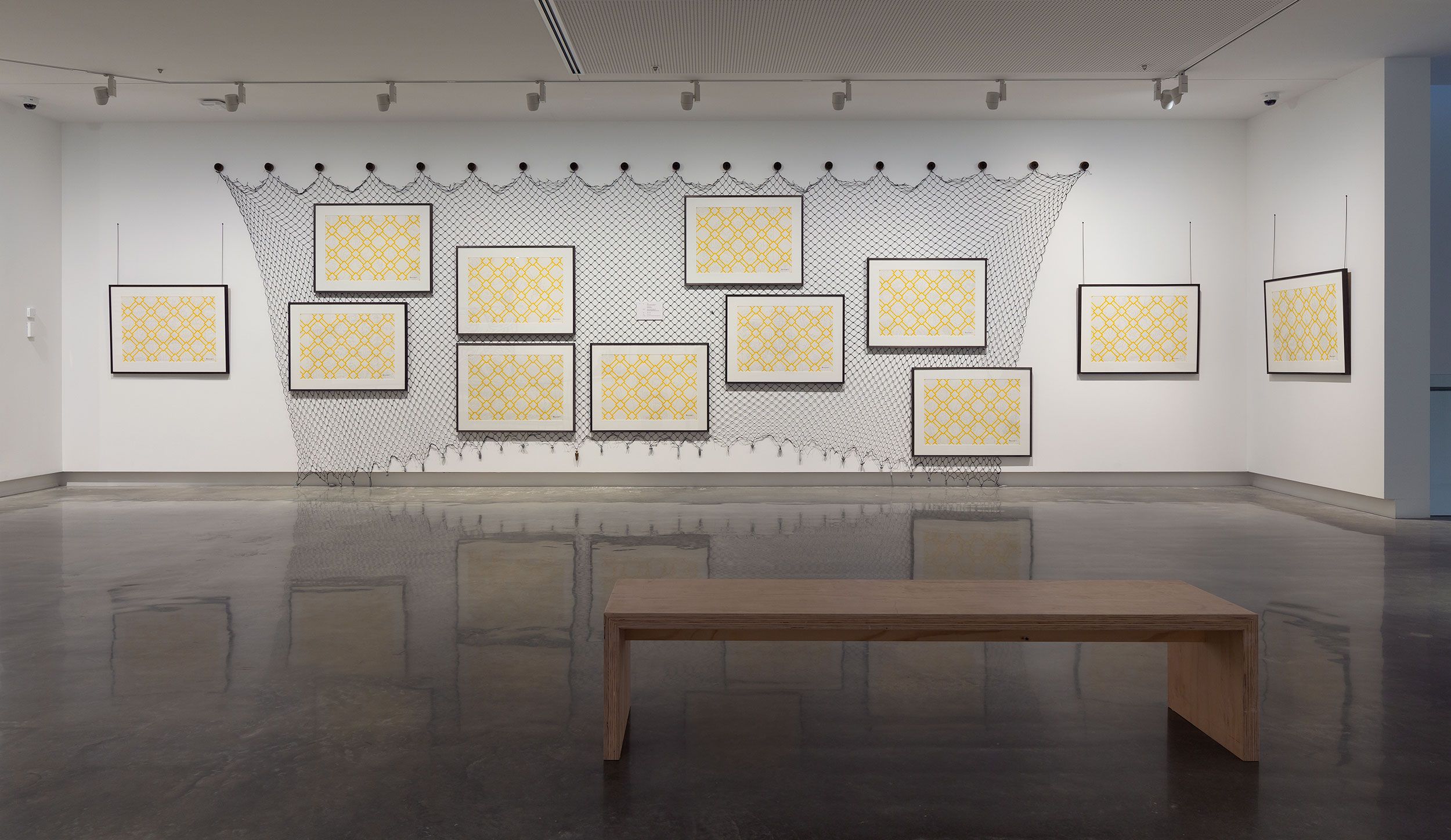 A gallery wall displays twelve framed geometric yellow artworks arranged in front of a net-like installation. The floor is polished, and a simple wooden bench sits in the foreground.