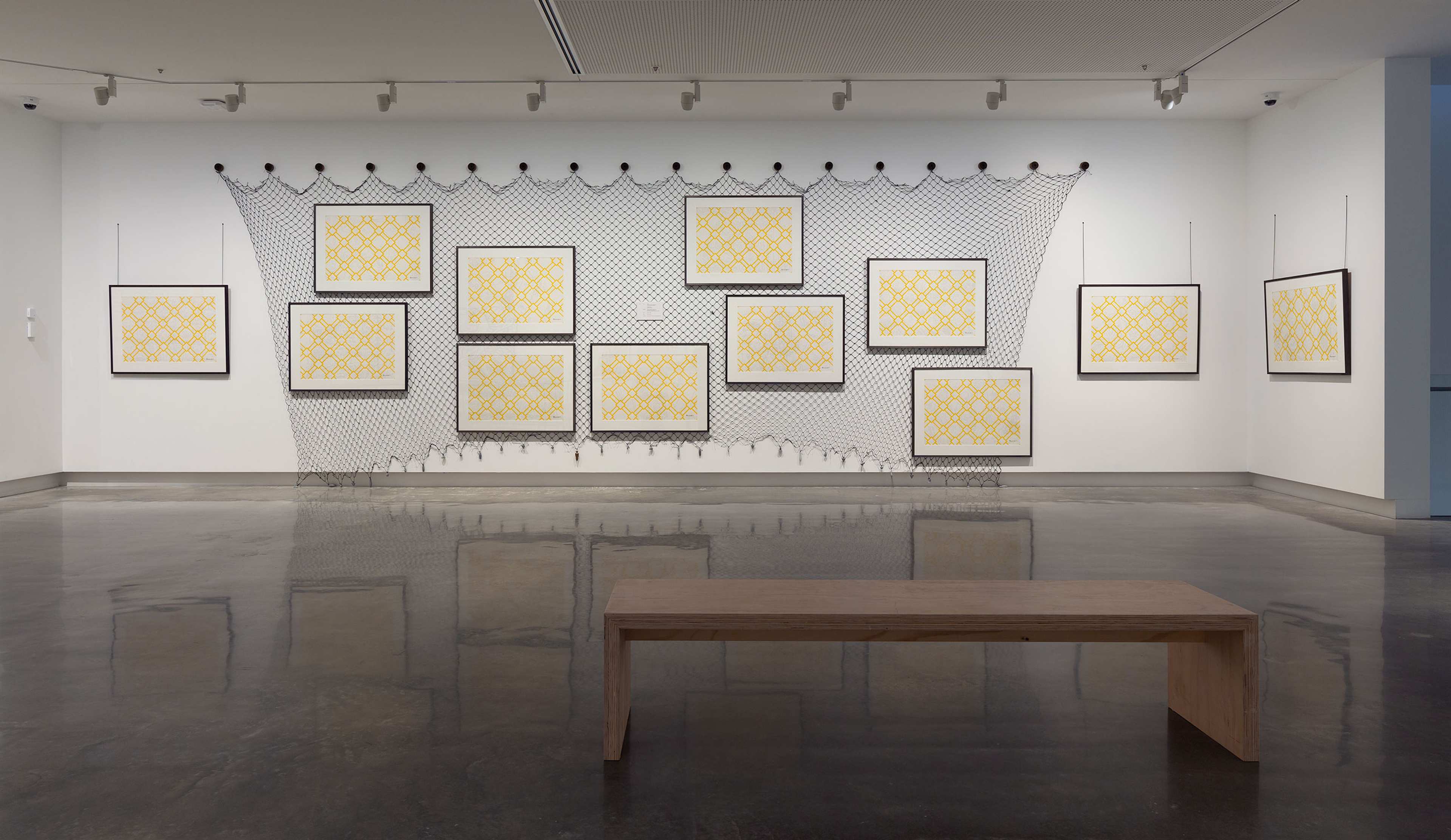 A gallery wall displays twelve framed geometric yellow artworks arranged in front of a net-like installation. The floor is polished, and a simple wooden bench sits in the foreground.