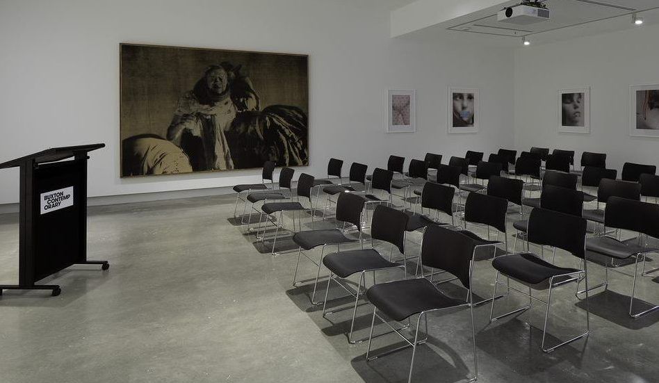 A modern, empty lecture room with rows of black chairs facing a podium on the left. The walls display framed photographs and a large, dark-toned painting. The floor is concrete and the space is well-lit.