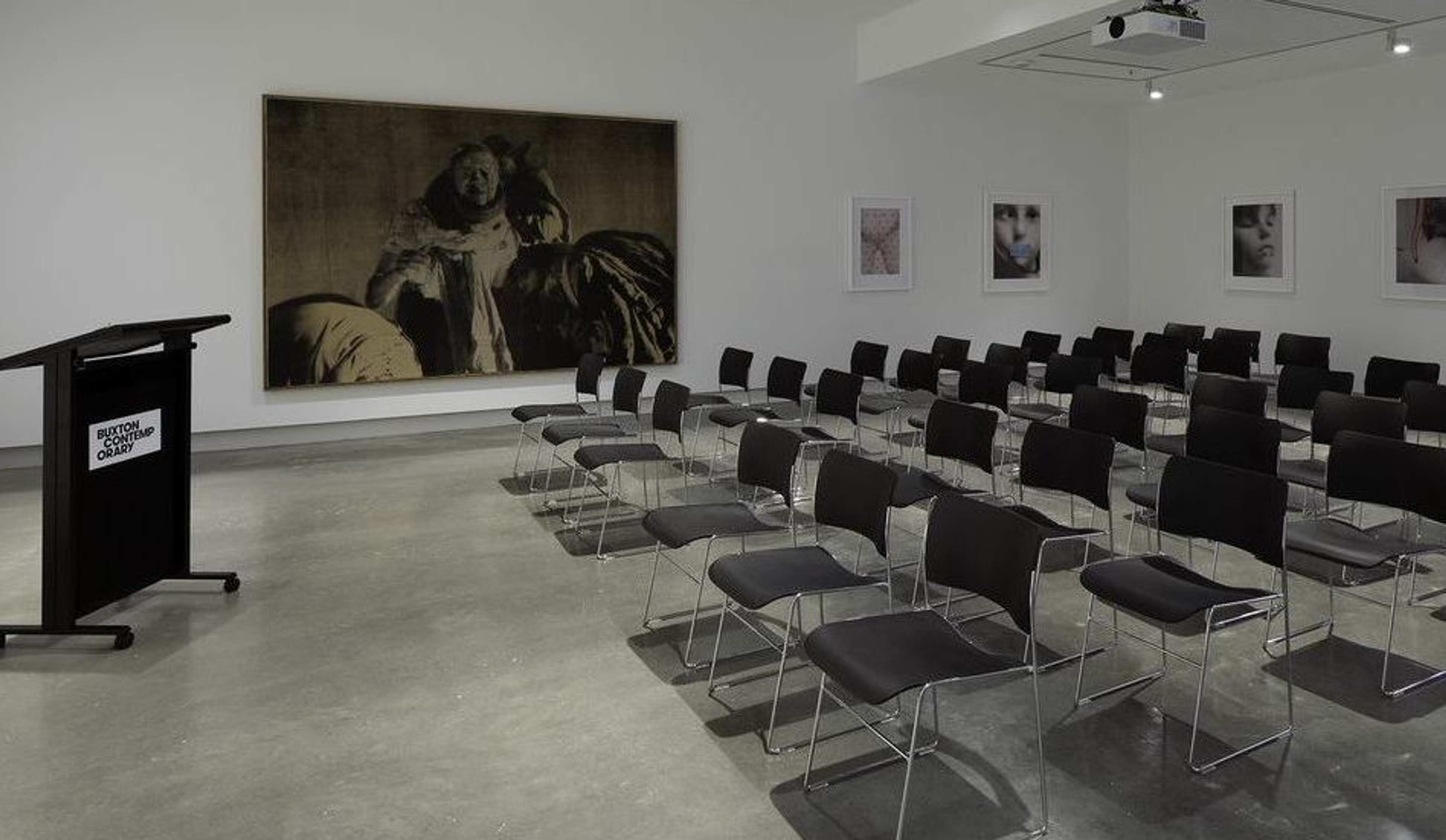 A modern, empty lecture room with rows of black chairs facing a podium on the left. The walls display framed photographs and a large, dark-toned painting. The floor is concrete and the space is well-lit.