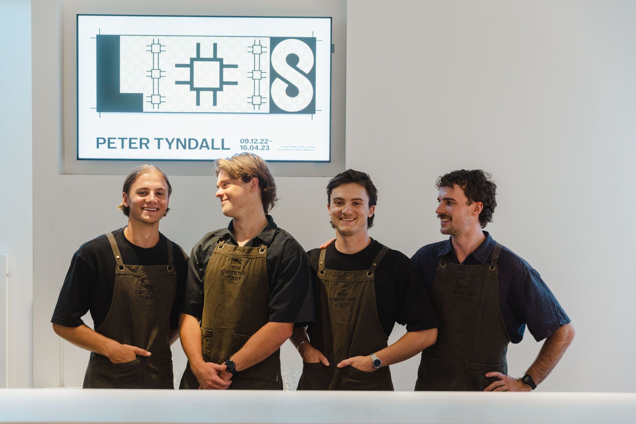 Four waiters with dark aprons stand against a white wall with a sign saying Peter Tyndall