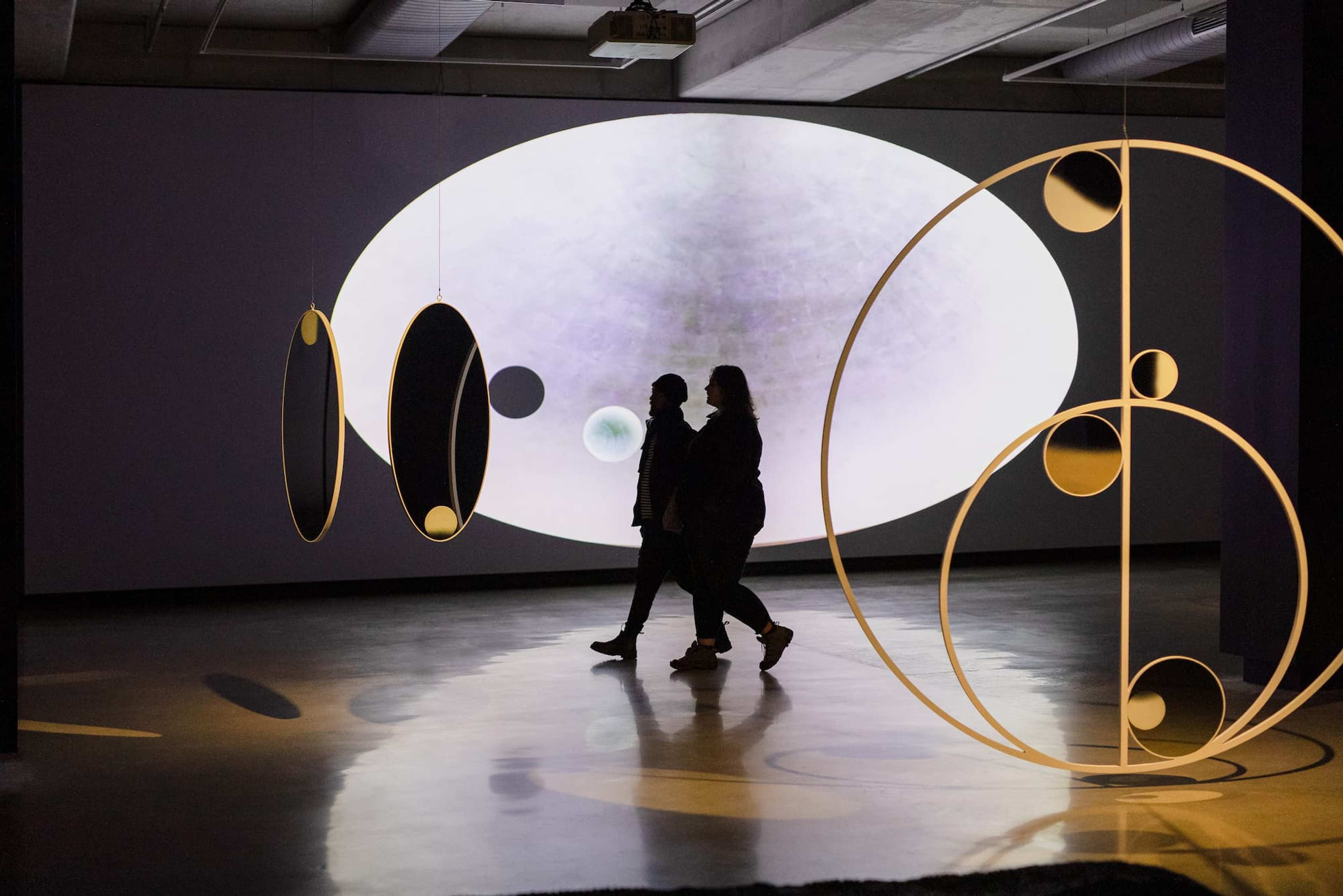 Two people are silhouetted as they walk past large circular and oval art installations, with a glowing oval light and abstract shapes projected on the wall behind them in a modern indoor space.