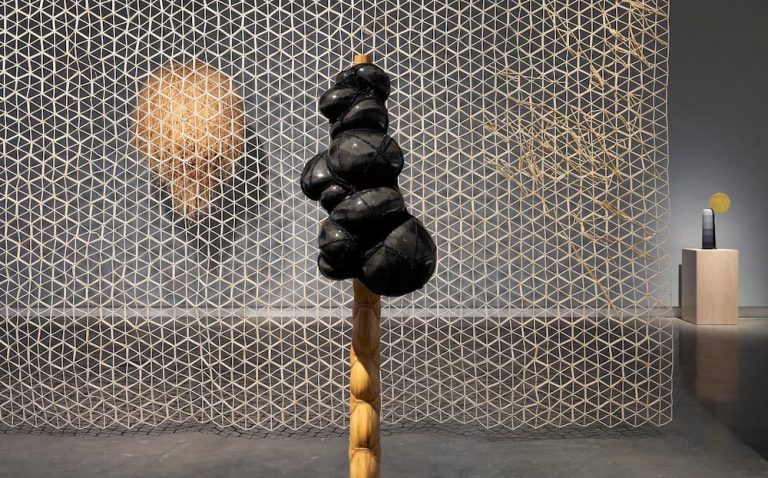 A modern art gallery features a geometric mesh backdrop with a woven circular piece, a black abstract sculpture on a twisted wooden base in the foreground, and a small pedestal with objects in the background.