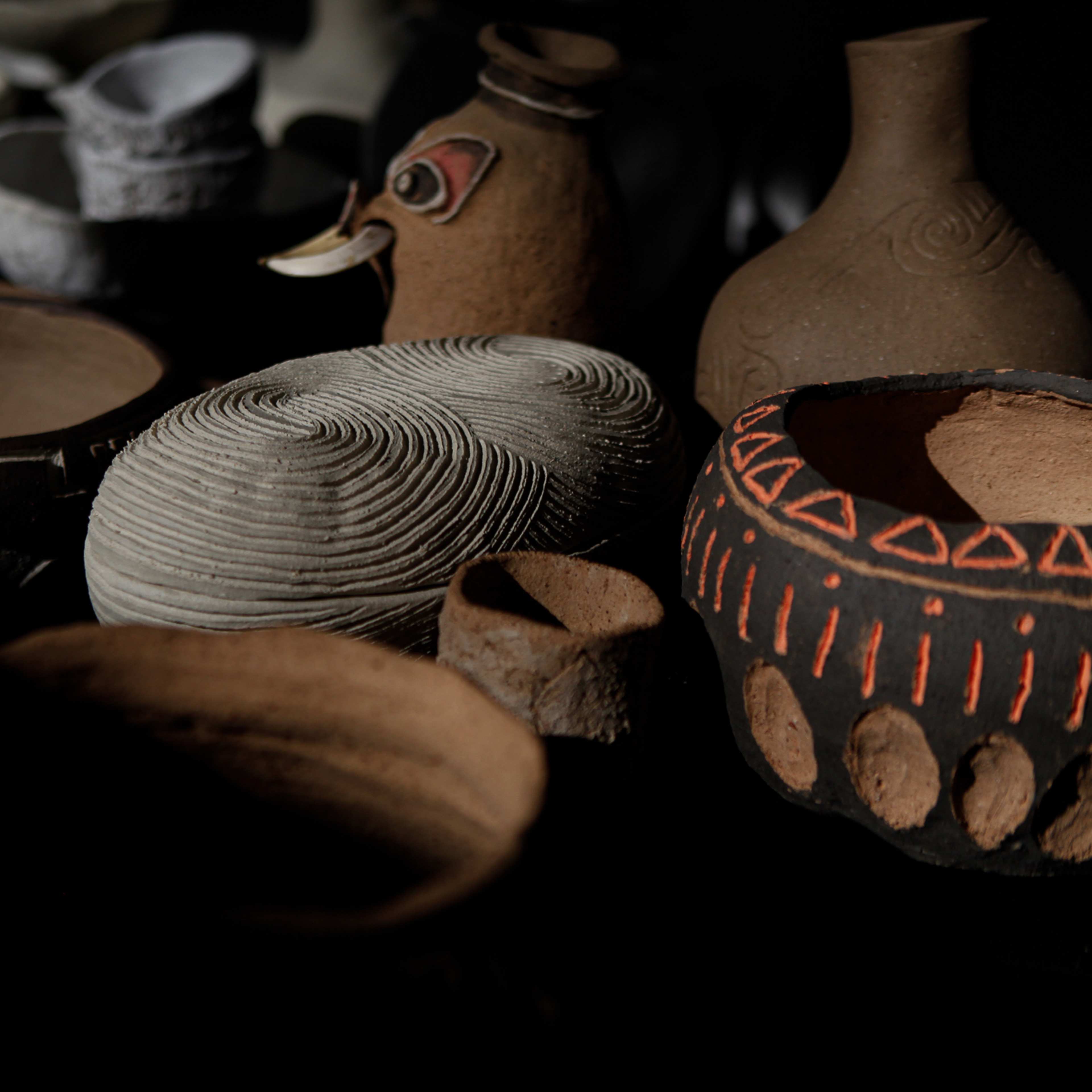 A collection of handmade pottery, including bowls, vases, and a decorative vessel with a beak-like spout, displayed in low light with intricate textures and patterns visible on the surfaces.