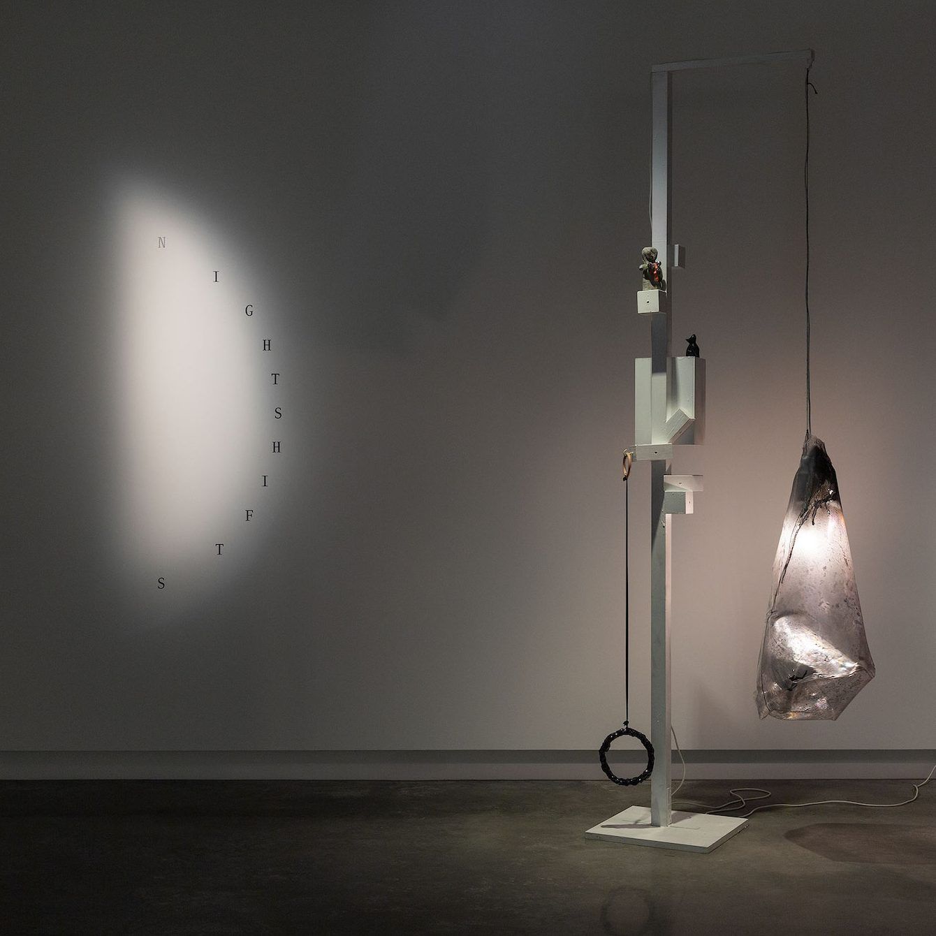 A modern art installation features a large, illuminated, translucent sculpture hanging from a white geometric stand. On the wall, the word NIGHTSHIFTS is spelled vertically in spaced-out letters. The setting is dimly lit.