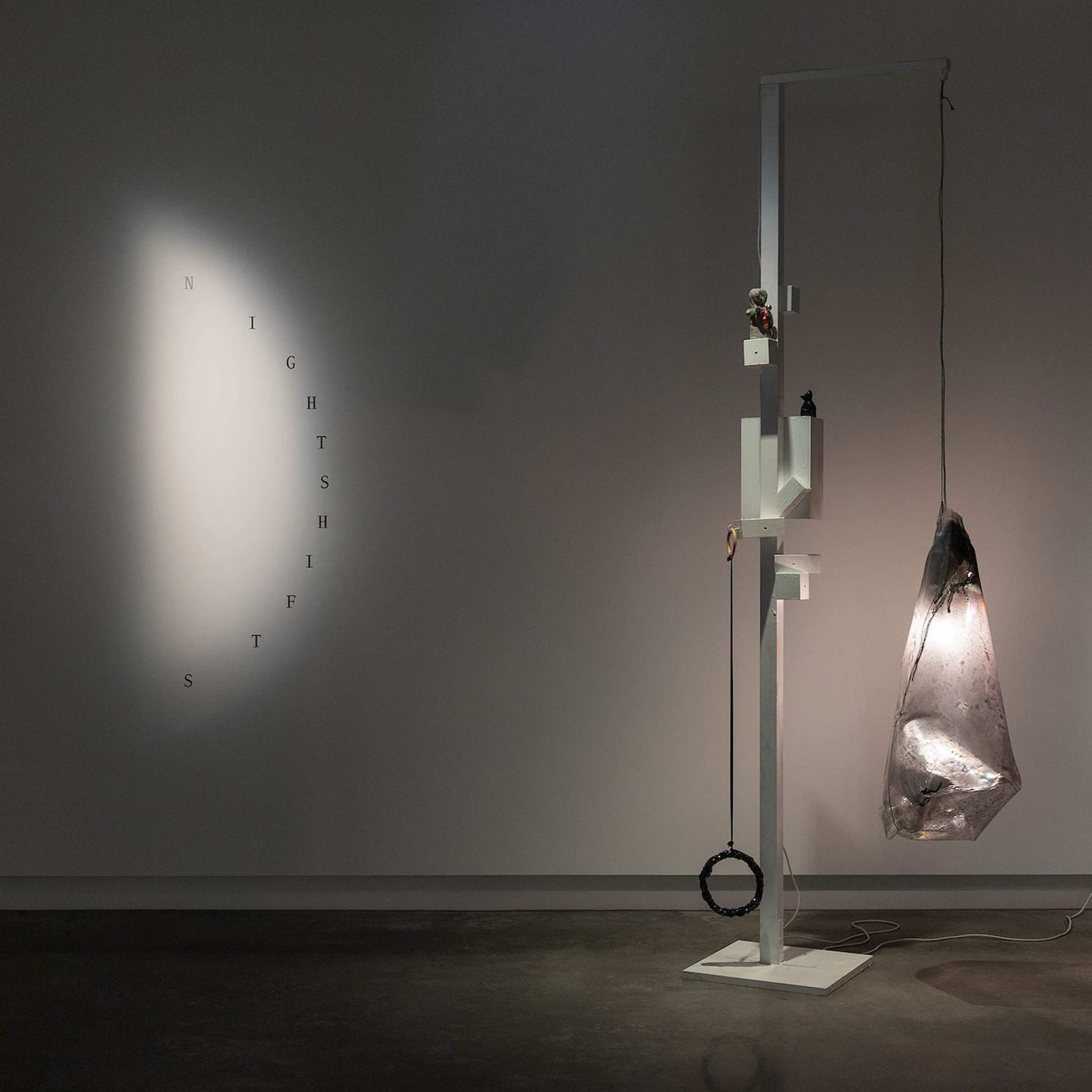 A modern art installation features a large, illuminated, translucent sculpture hanging from a white geometric stand. On the wall, the word NIGHTSHIFTS is spelled vertically in spaced-out letters. The setting is dimly lit.