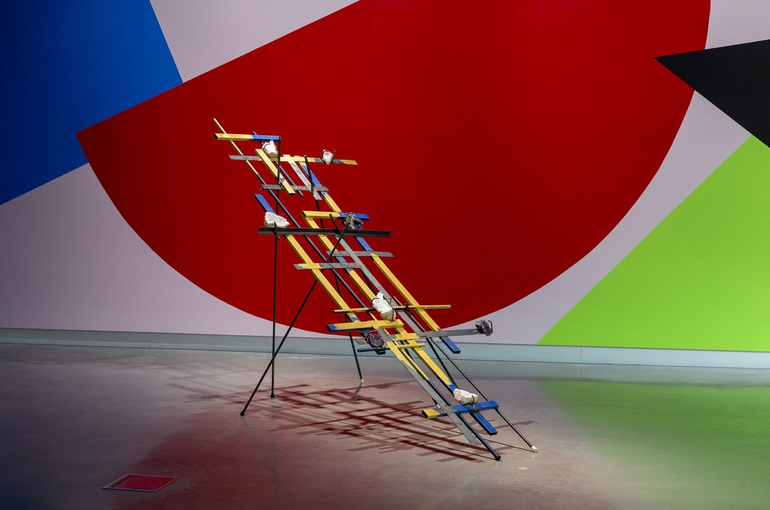 A geometric sculpture of interlaced blue and yellow wooden planks stands on metal legs, decorated with white objects, against a wall painted with large red, green, blue, and black angular shapes.
