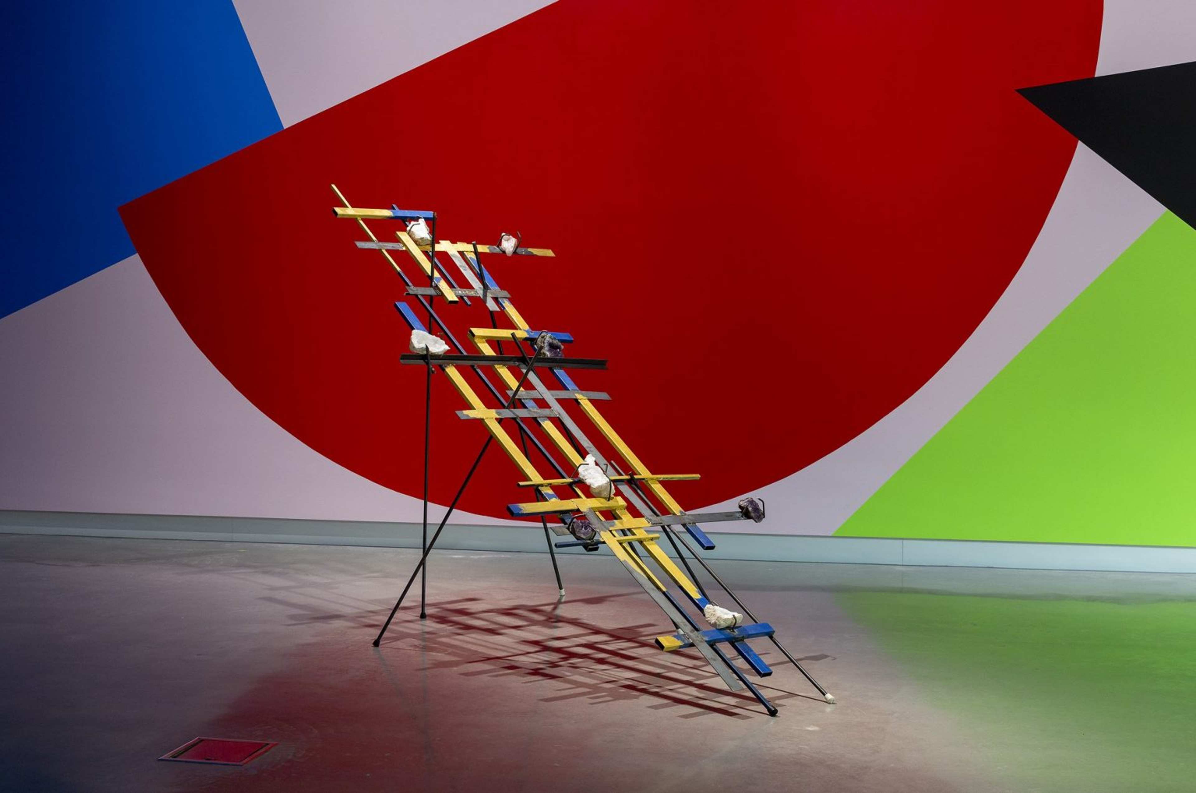 A geometric sculpture of interlaced blue and yellow wooden planks stands on metal legs, decorated with white objects, against a wall painted with large red, green, blue, and black angular shapes.