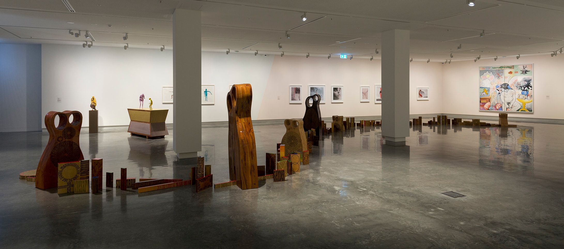 A contemporary art gallery with sculptures made of wood and mixed materials arranged along the floor, framed artwork on white walls, polished concrete floors, and bright ceiling lights.