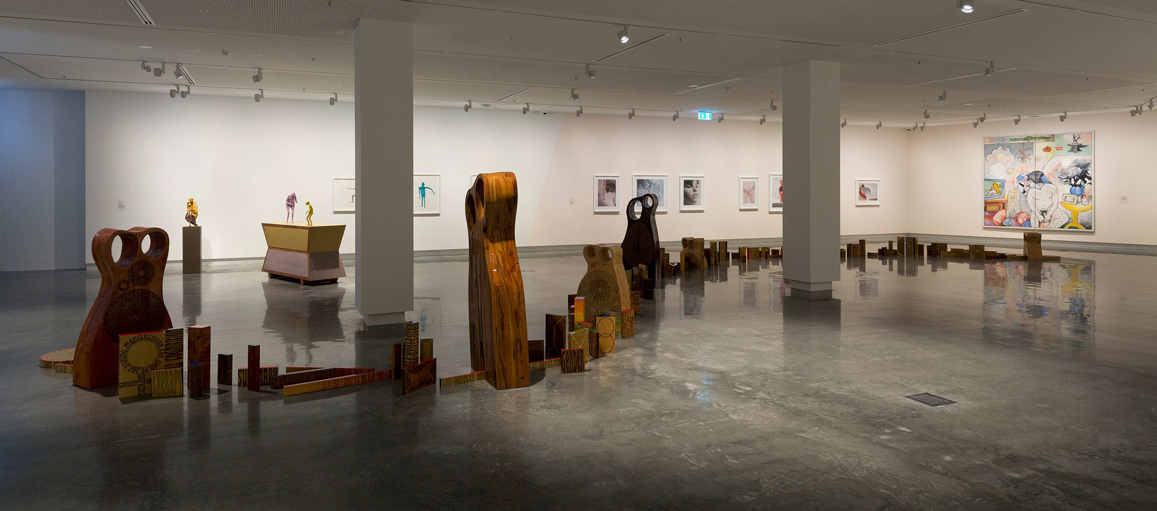 A contemporary art gallery with sculptures made of wood and mixed materials arranged along the floor, framed artwork on white walls, polished concrete floors, and bright ceiling lights.