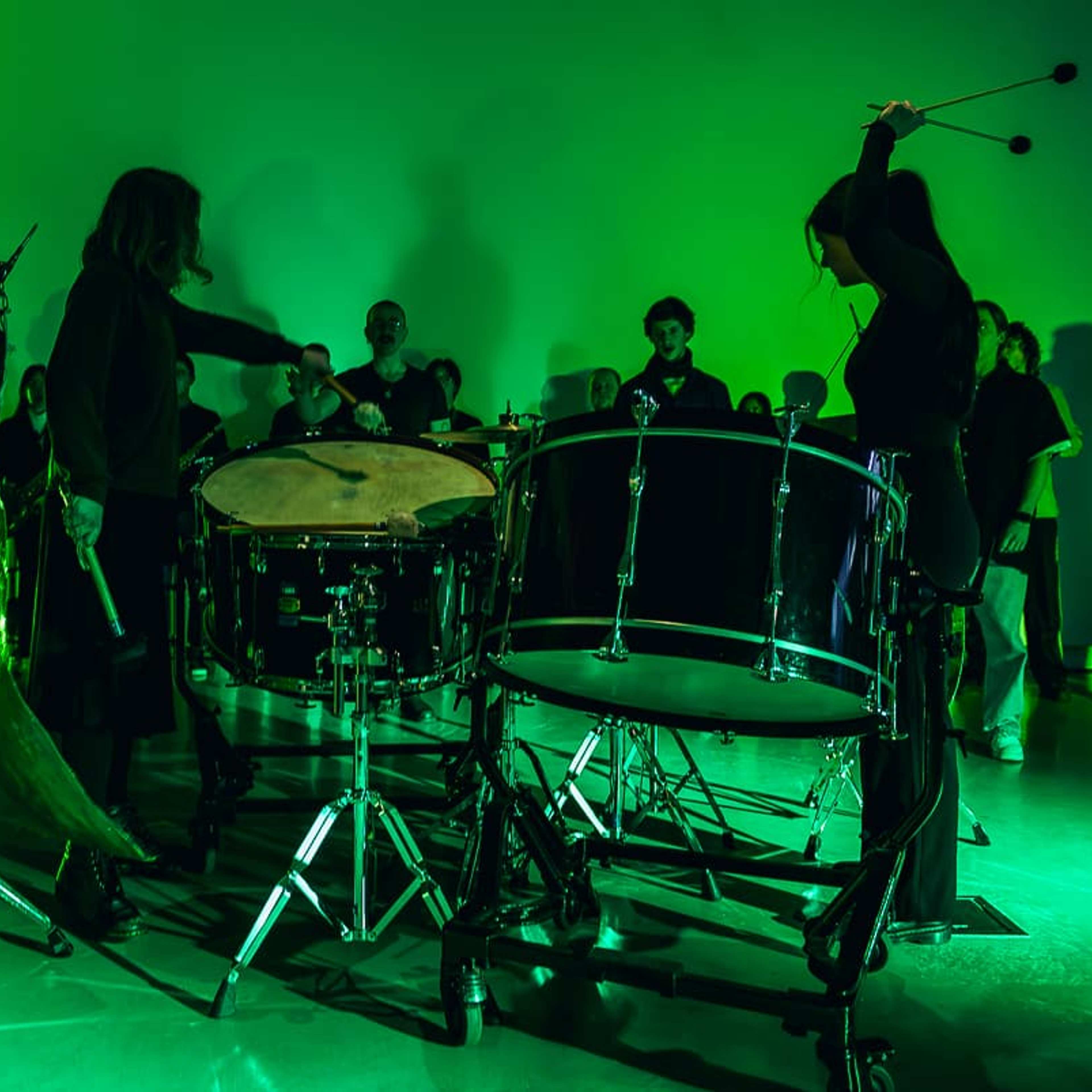 Musicians perform with large drums and percussion instruments on a stage lit by green light, with a group of people standing in the background watching the performance.