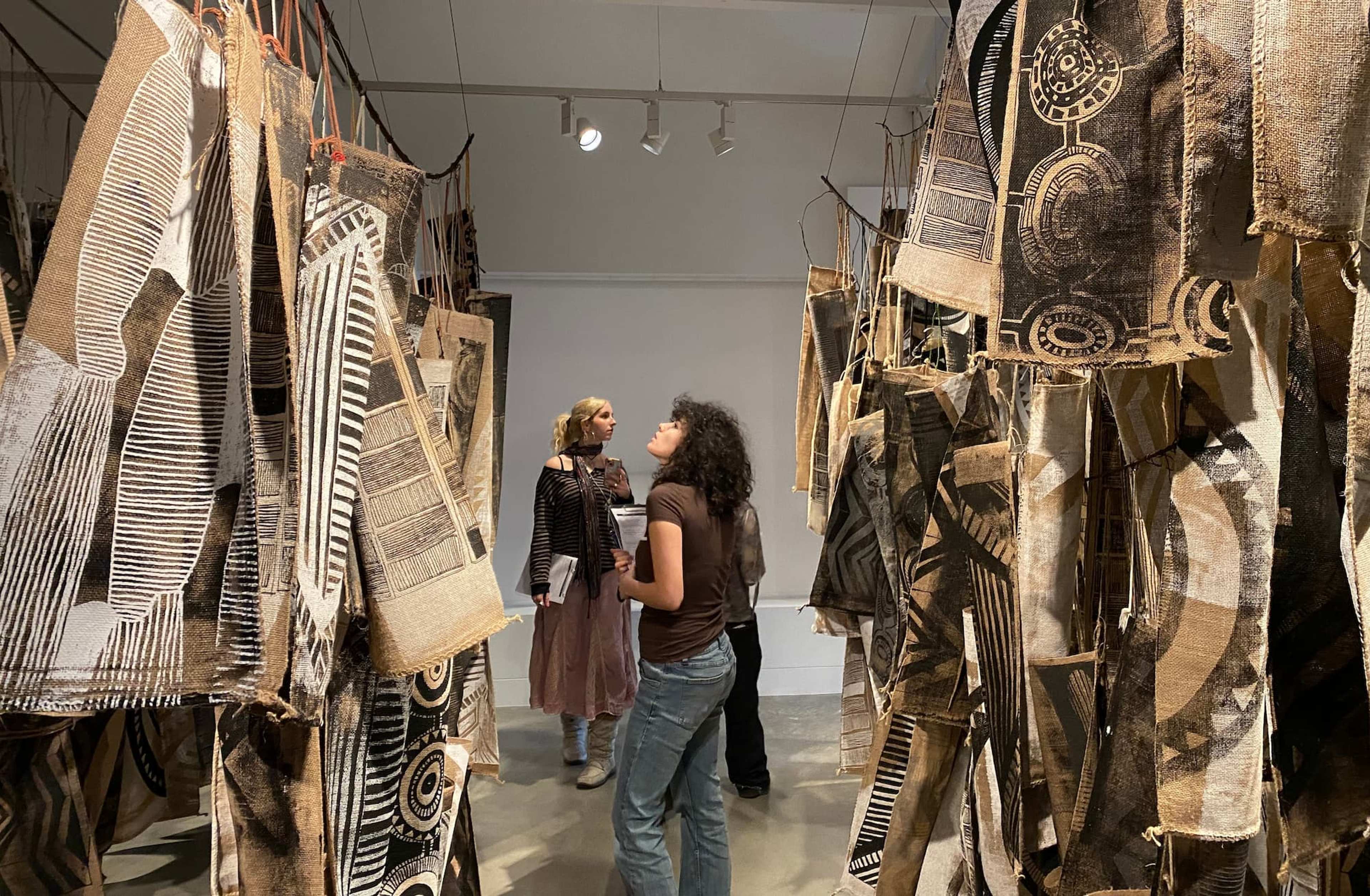 Two people stand in a gallery, observing hanging fabric artworks with geometric and abstract patterns in brown, black, and white tones. Spotlights above illuminate the textured art pieces.
