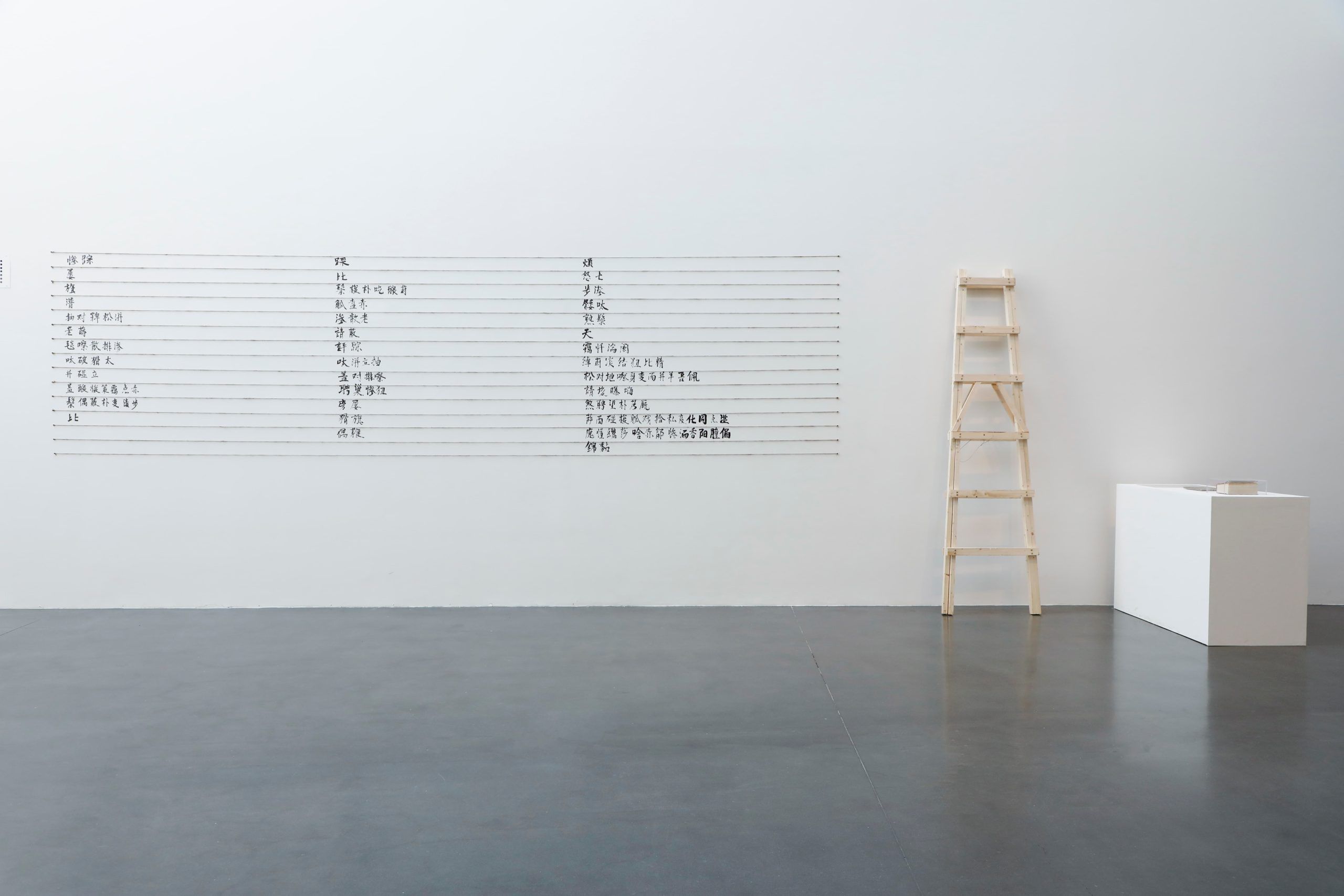 A minimalist gallery space with a wooden ladder, a white pedestal displaying an object, and a large chart with rows of text in a Chinese script mounted on a white wall. The floor is polished concrete.