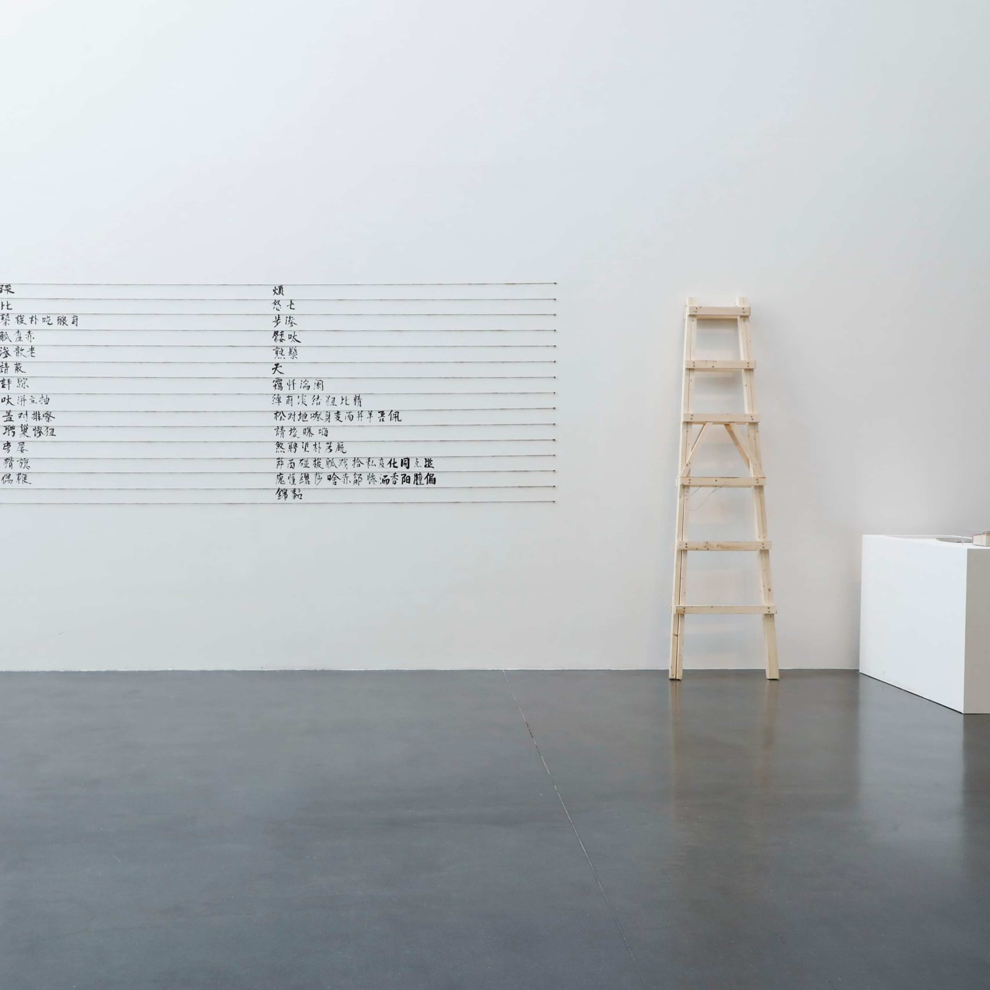A minimalist gallery space with a wooden ladder, a white pedestal displaying an object, and a large chart with rows of text in a Chinese script mounted on a white wall. The floor is polished concrete.
