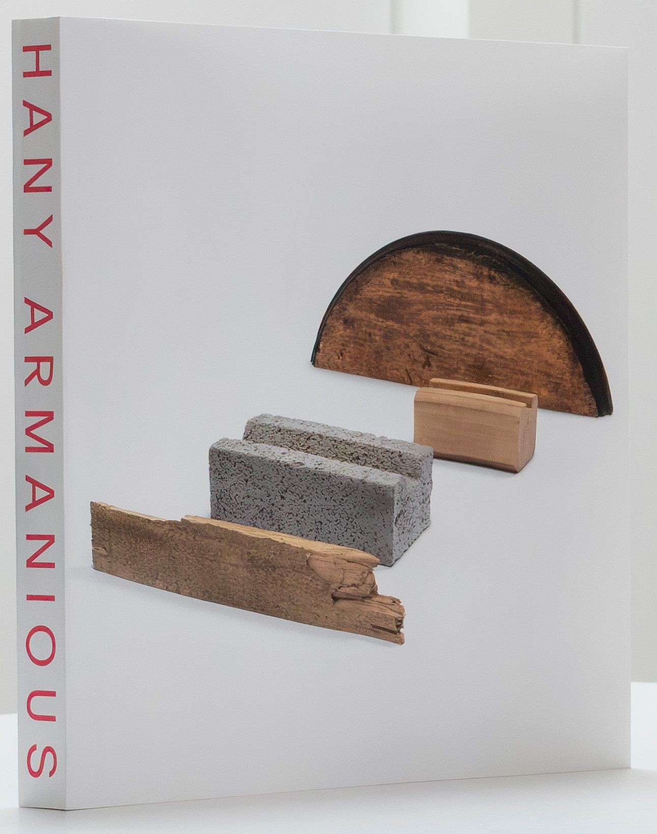 A book with HANY ARMANIOUS in red text on the spine. The cover features an arrangement of varied materials: a curved wooden piece, a gray cinder block, a rectangular wood block, and a strip of rough wood.