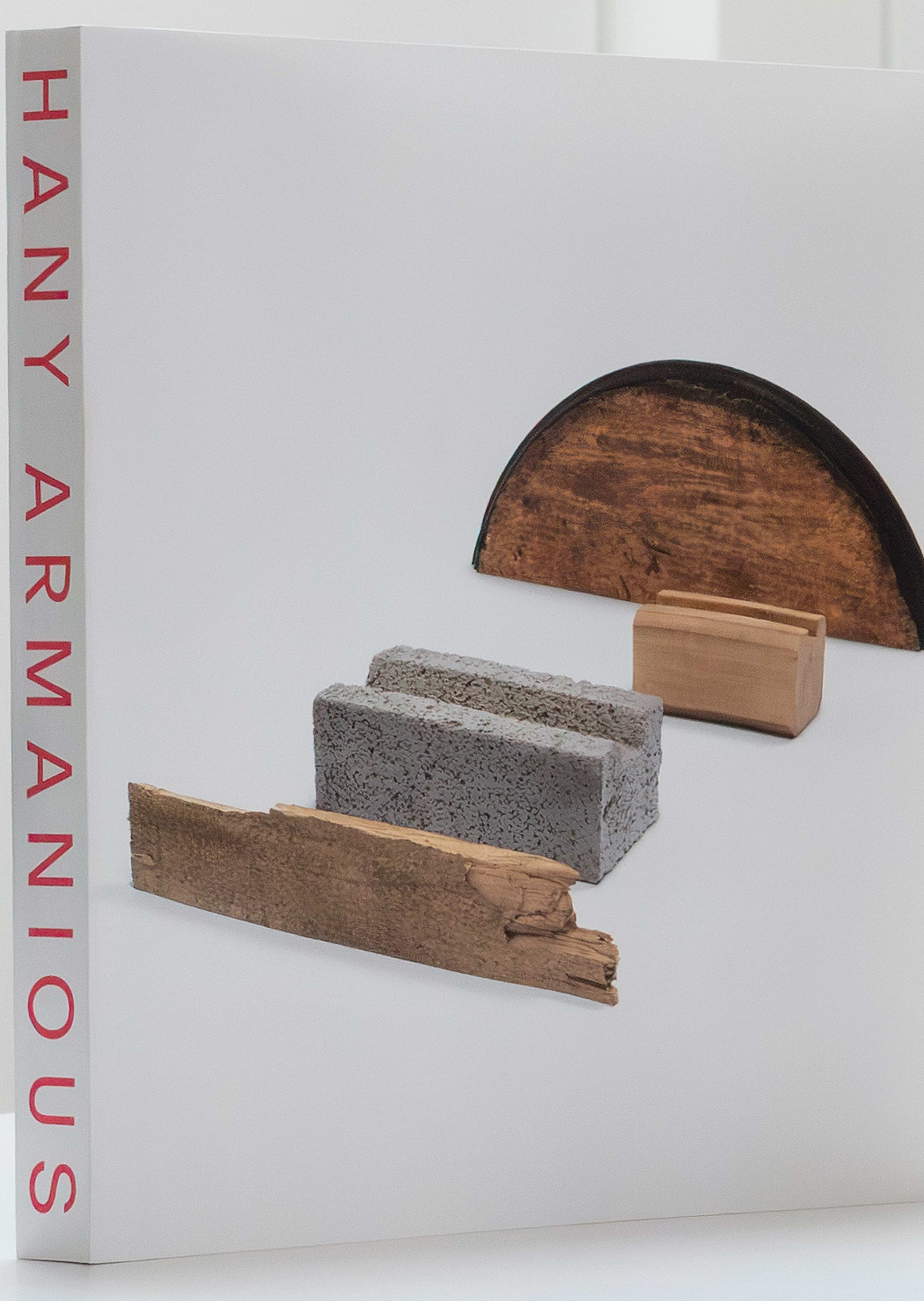 A book with HANY ARMANIOUS in red text on the spine. The cover features an arrangement of varied materials: a curved wooden piece, a gray cinder block, a rectangular wood block, and a strip of rough wood.