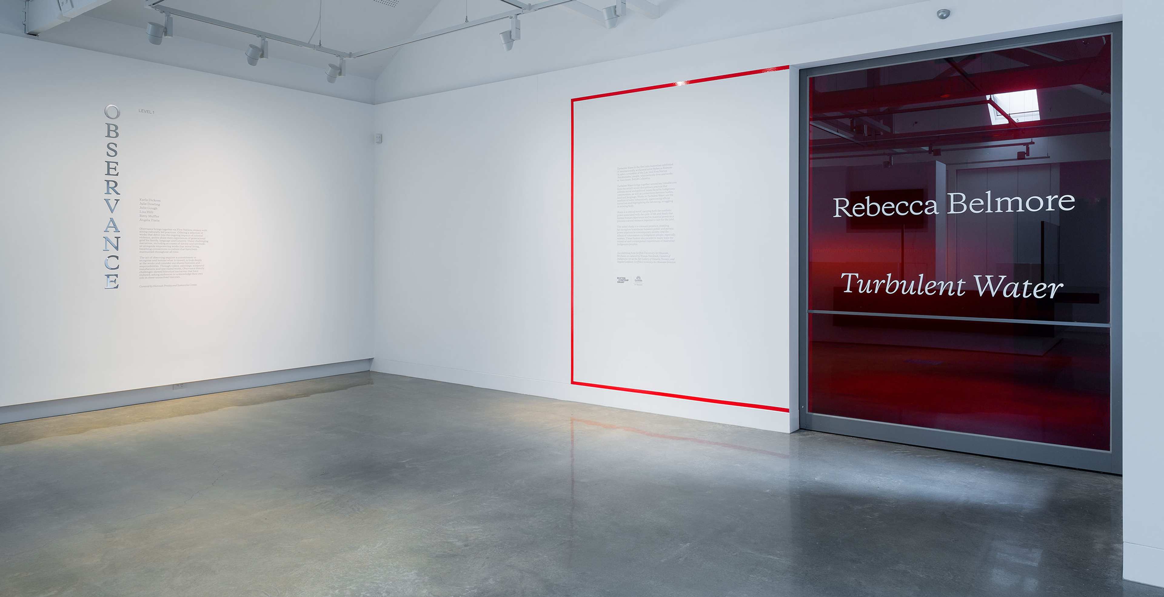 A modern gallery space with white walls, grey concrete floors, and track lighting. On the left wall is the word Observance vertically. A glass door on the right reads Rebecca Belmore Turbulent Water.
