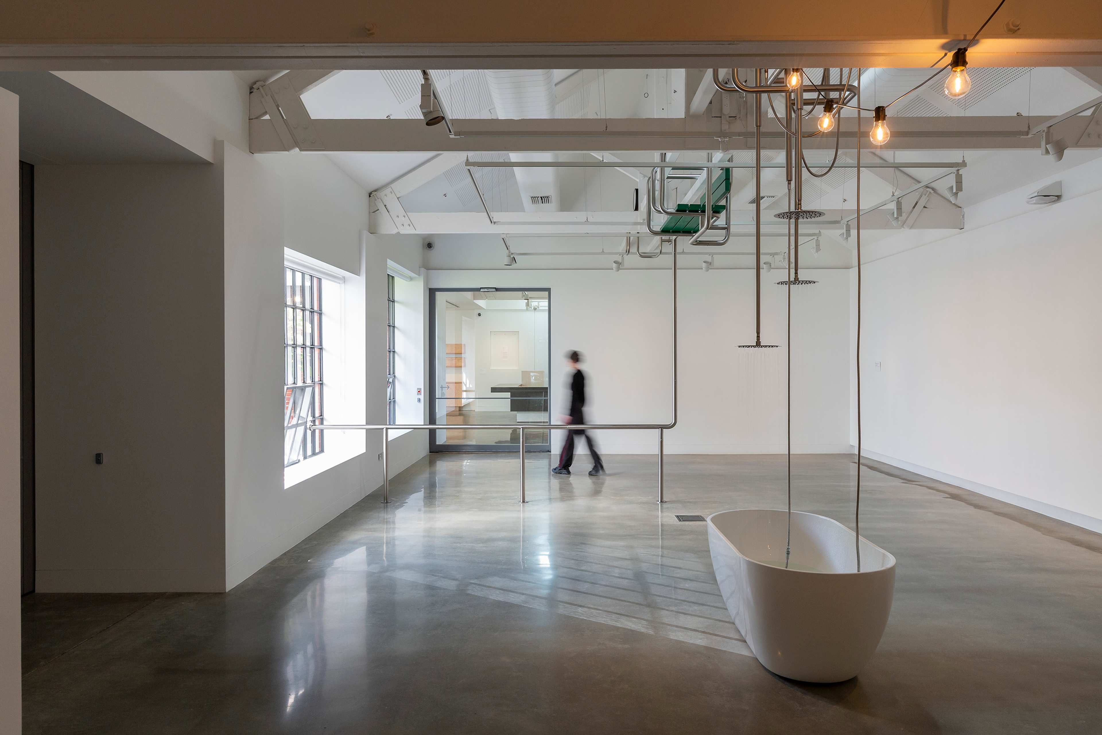 A modern, minimalistic room with polished concrete floors, white walls, a freestanding bathtub, and exposed ceiling pipes. Natural light enters through large windows; a person walks past a railing in the background.