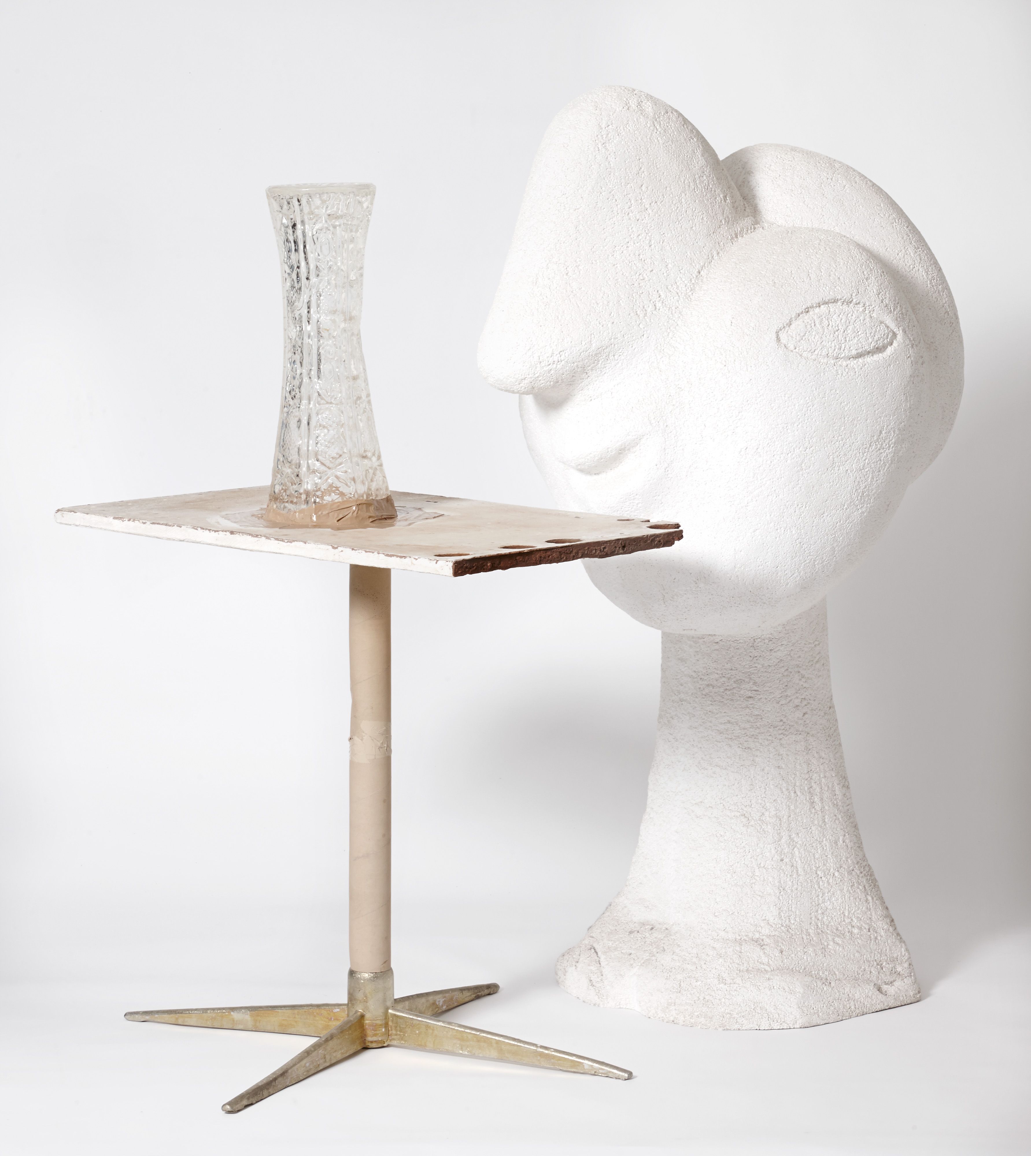 A clear vase on top of a single stand table, in front of a white sculpture.