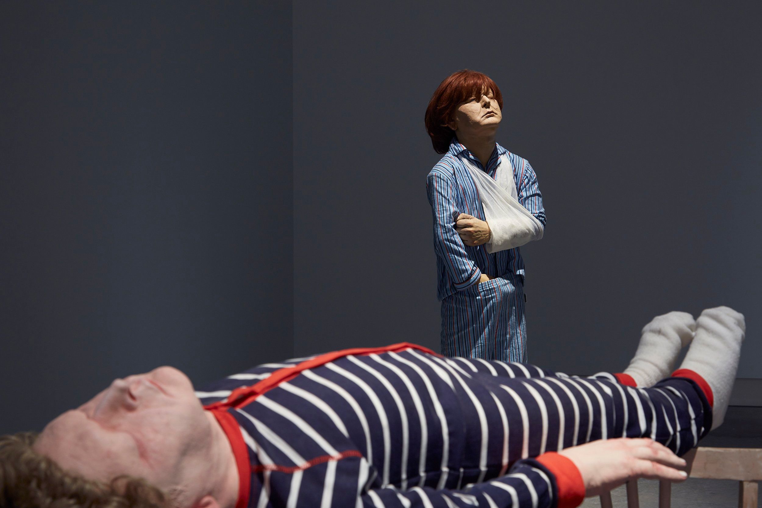 Two realistic, life-sized sculptures in a dimly lit room: one of a person lying down in striped pajamas, and another standing in blue pajamas with a sling on their arm, gazing upward with a solemn expression.