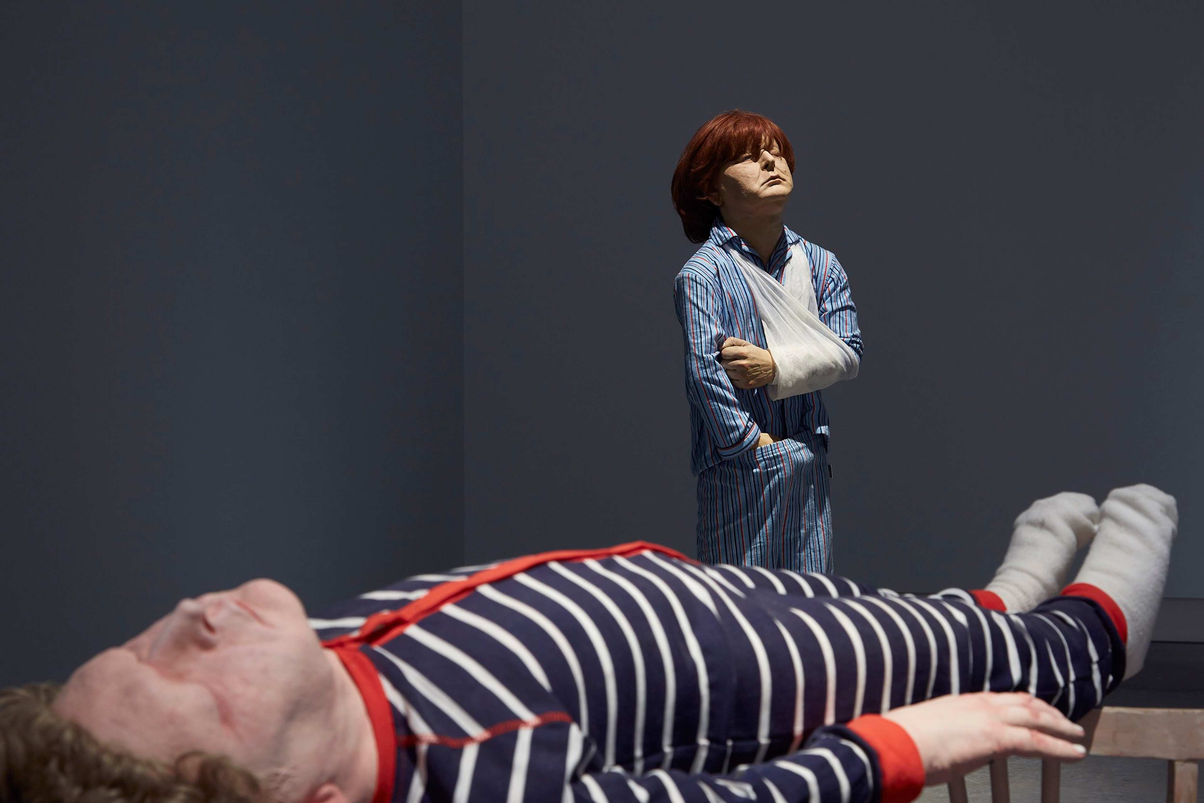 Two realistic, life-sized sculptures in a dimly lit room: one of a person lying down in striped pajamas, and another standing in blue pajamas with a sling on their arm, gazing upward with a solemn expression.