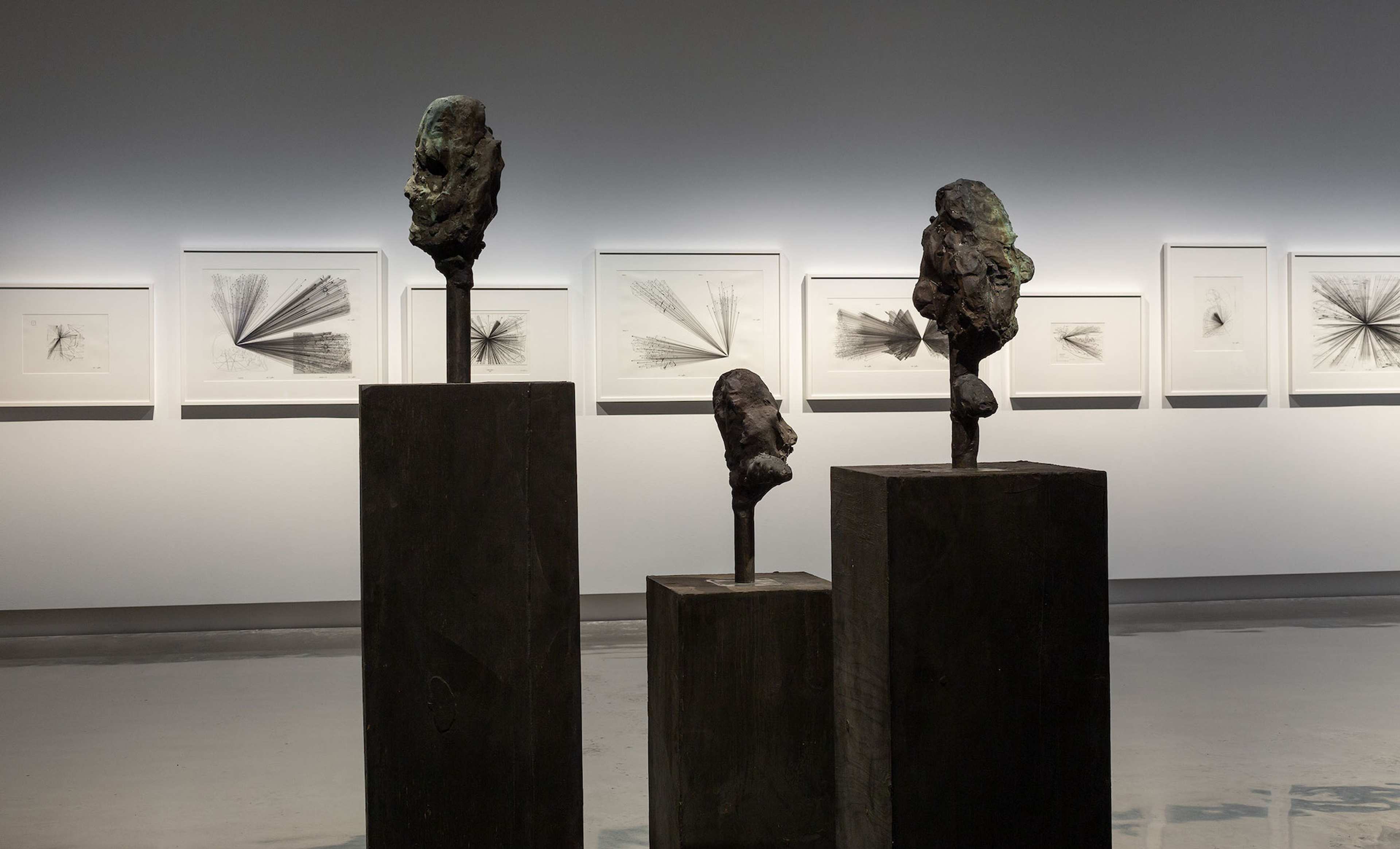 Three abstract bronze head sculptures are displayed on tall dark pedestals in a gallery, with black-and-white geometric drawings framed on the wall behind them, illuminated by soft lighting.
