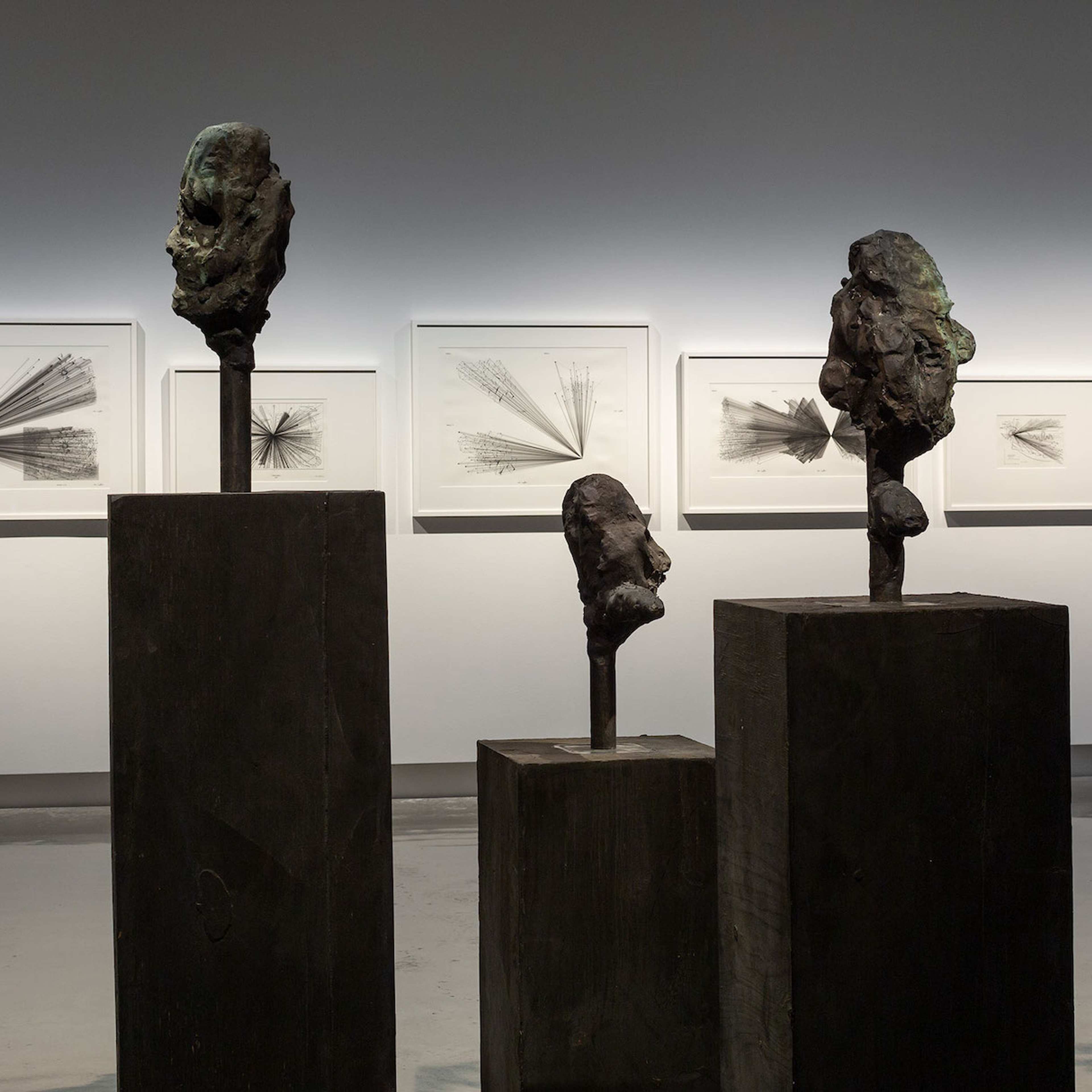 Three abstract bronze head sculptures are displayed on tall dark pedestals in a gallery, with black-and-white geometric drawings framed on the wall behind them, illuminated by soft lighting.