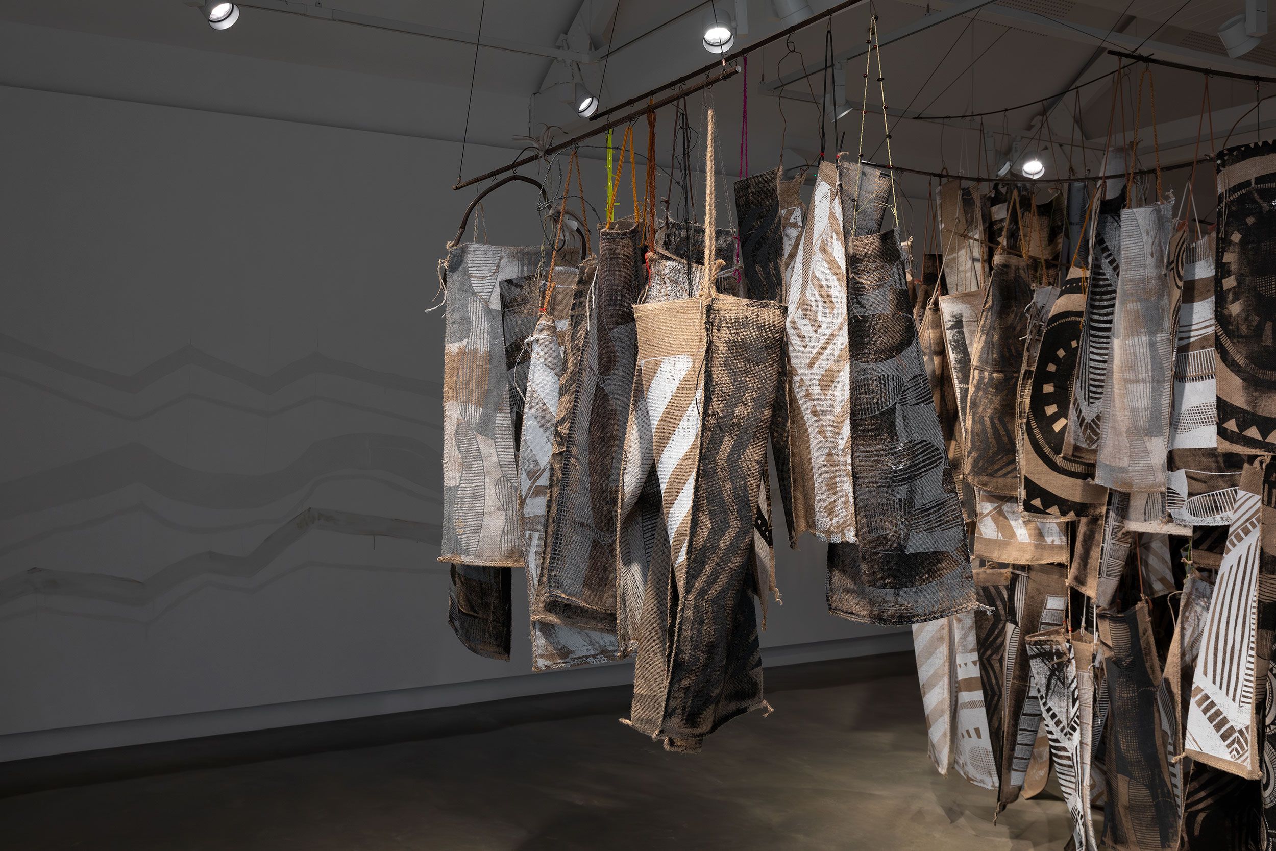 Fabric panels with abstract black and white patterns hang from rods in an art gallery. Spotlights illuminate the textured textiles, while shadows and wavy lines appear on the gray wall in the background.