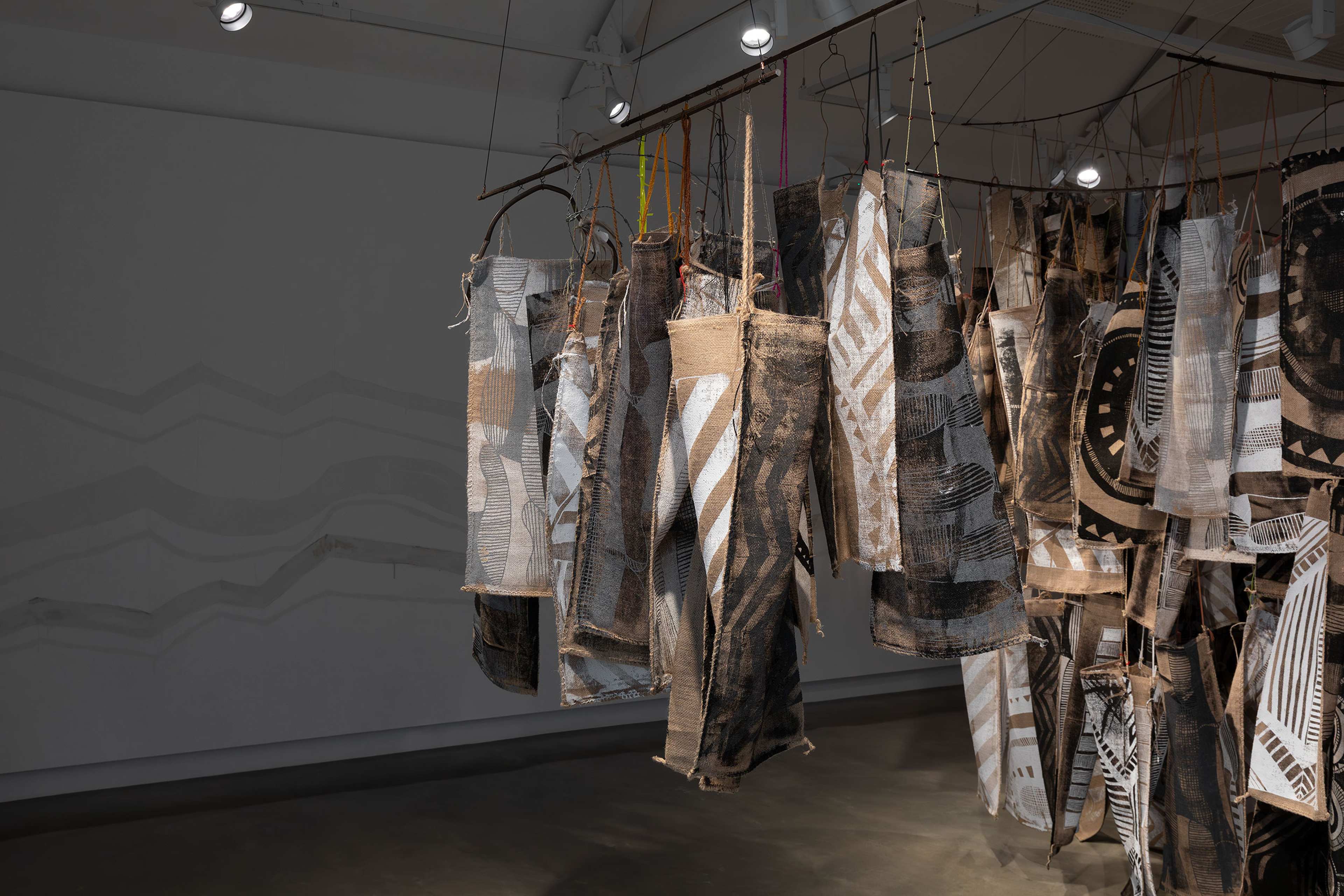 Fabric panels with abstract black and white patterns hang from rods in an art gallery. Spotlights illuminate the textured textiles, while shadows and wavy lines appear on the gray wall in the background.