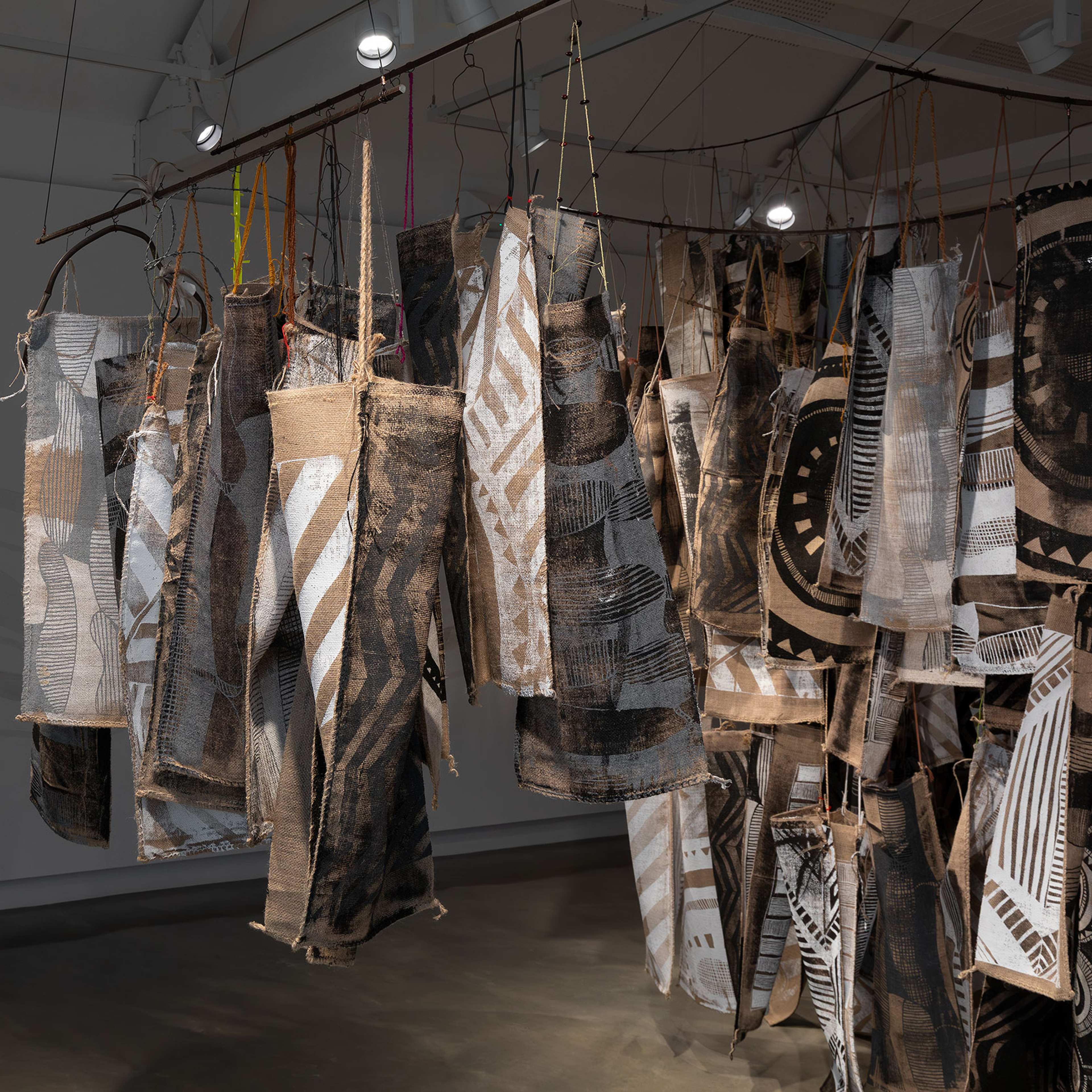 Fabric panels with abstract black and white patterns hang from rods in an art gallery. Spotlights illuminate the textured textiles, while shadows and wavy lines appear on the gray wall in the background.