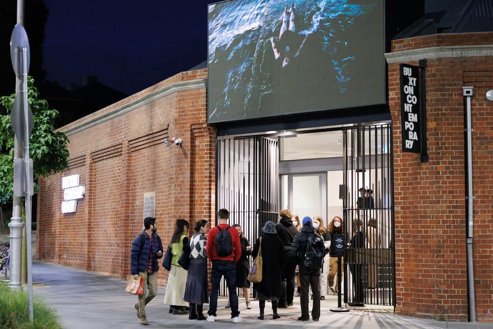 A group of people gather at the entrance of a contemporary art gallery in a brick building at night, with a large screen displaying artwork above the doorway.