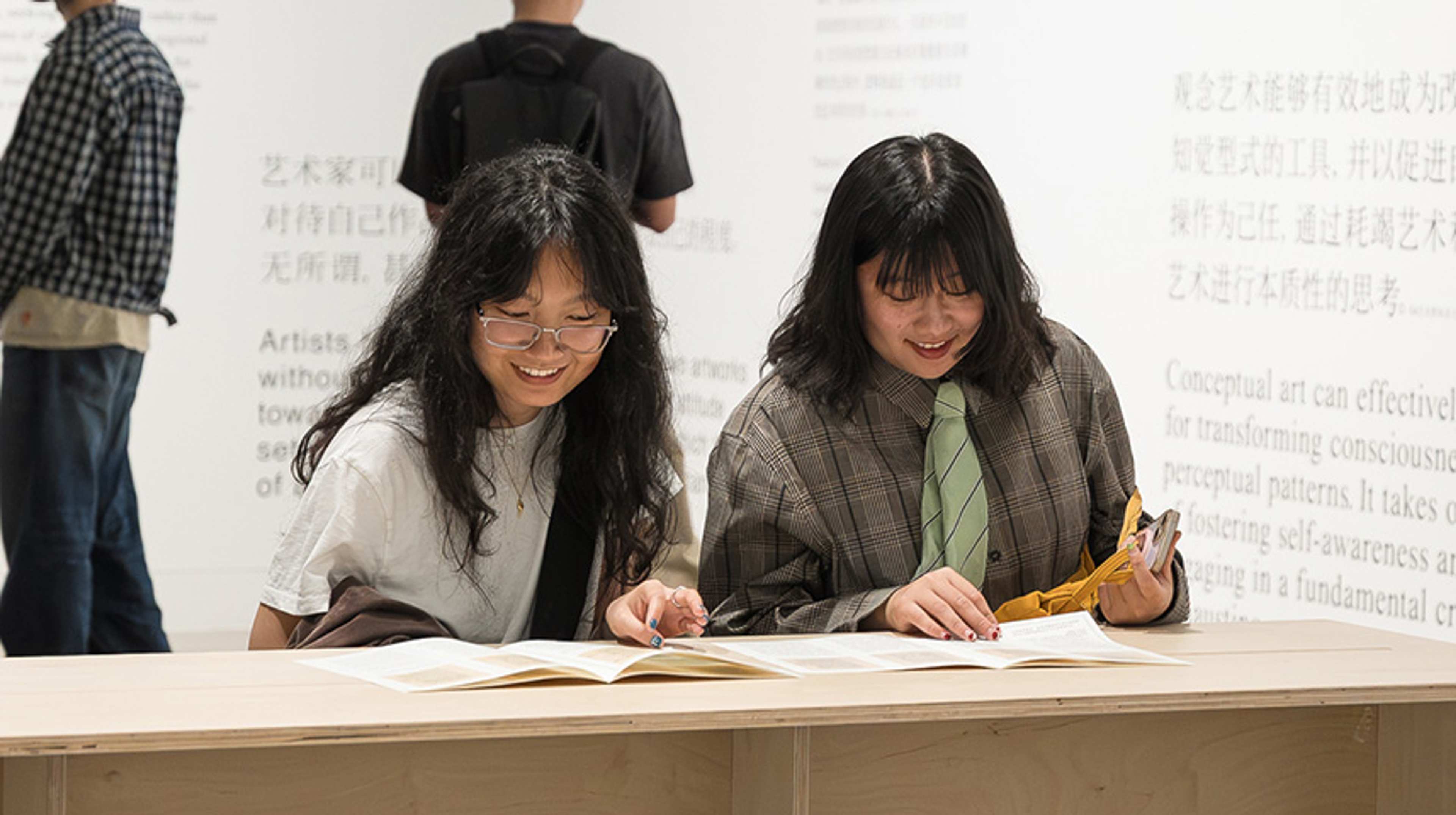 Two people sit at a table reading a magazing. Two people standing behind read chinese and englist text on a white wall in a contemporary art gallery