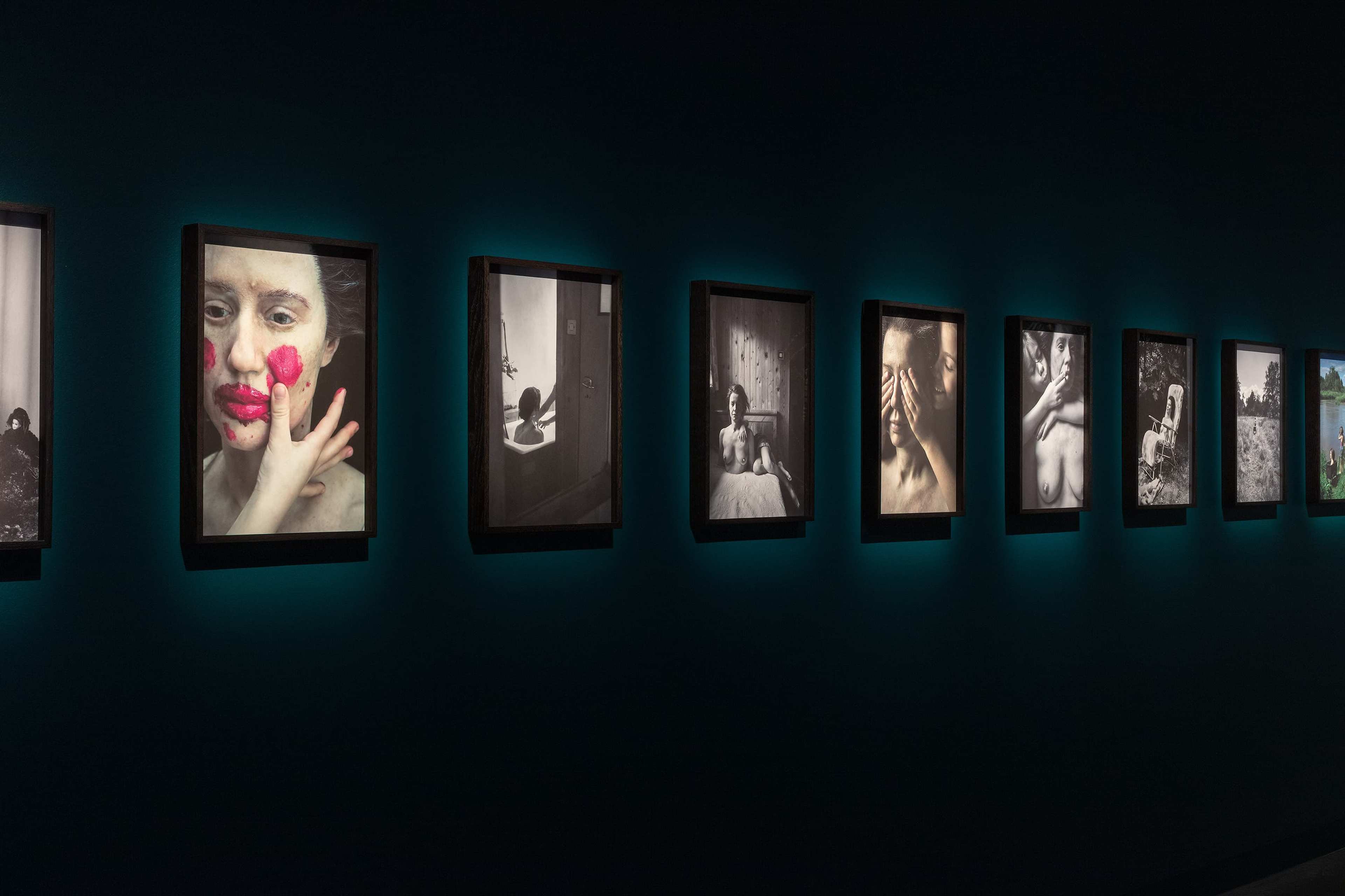 A row of framed black-and-white and color photographs displayed on a dark wall, each photo individually lit and featuring expressive portraits and artistic scenes.