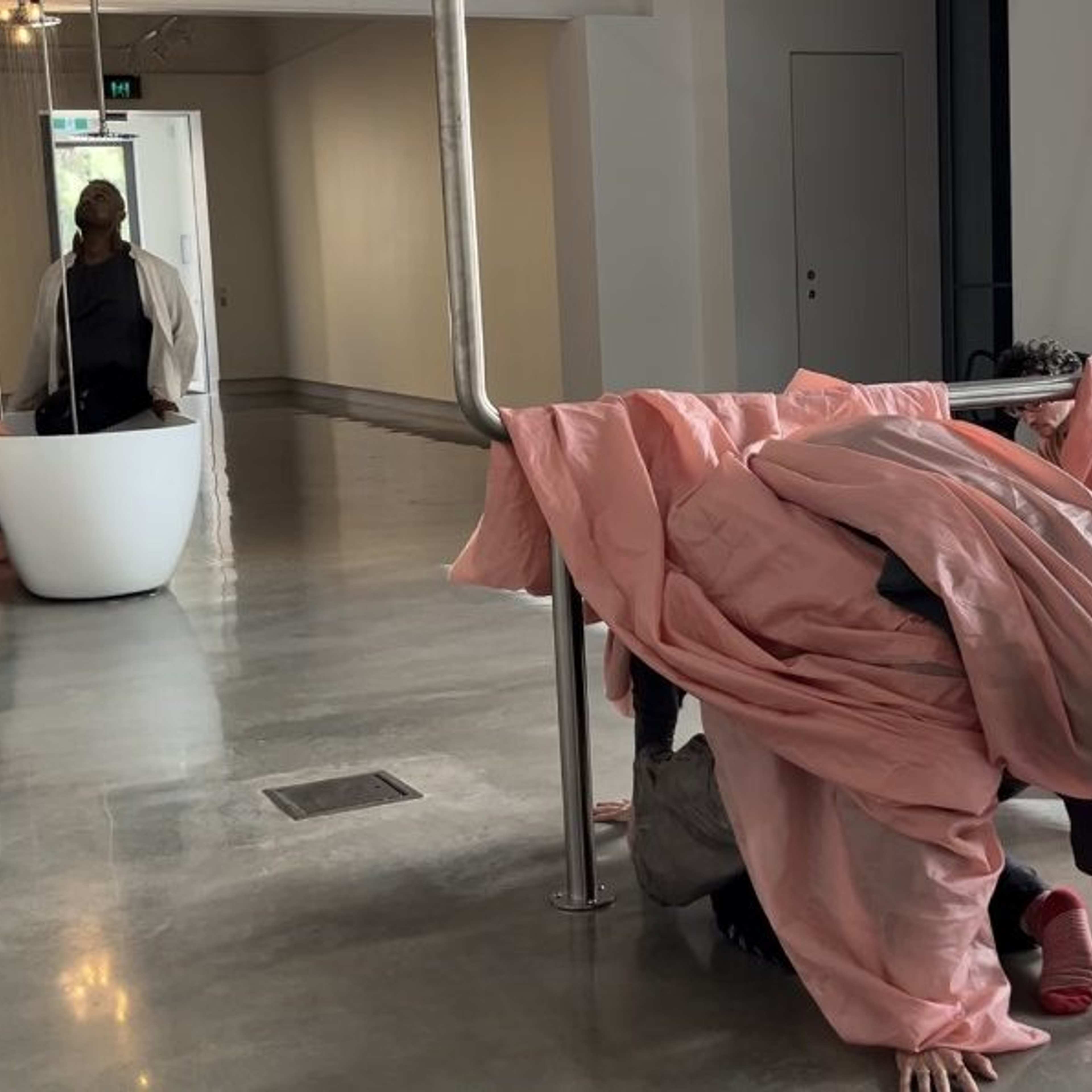 Several people in an art gallery interact with pink fabric draped over metal bars; one person crouches under the fabric, while others sit or move nearby on a shiny floor near large windows.