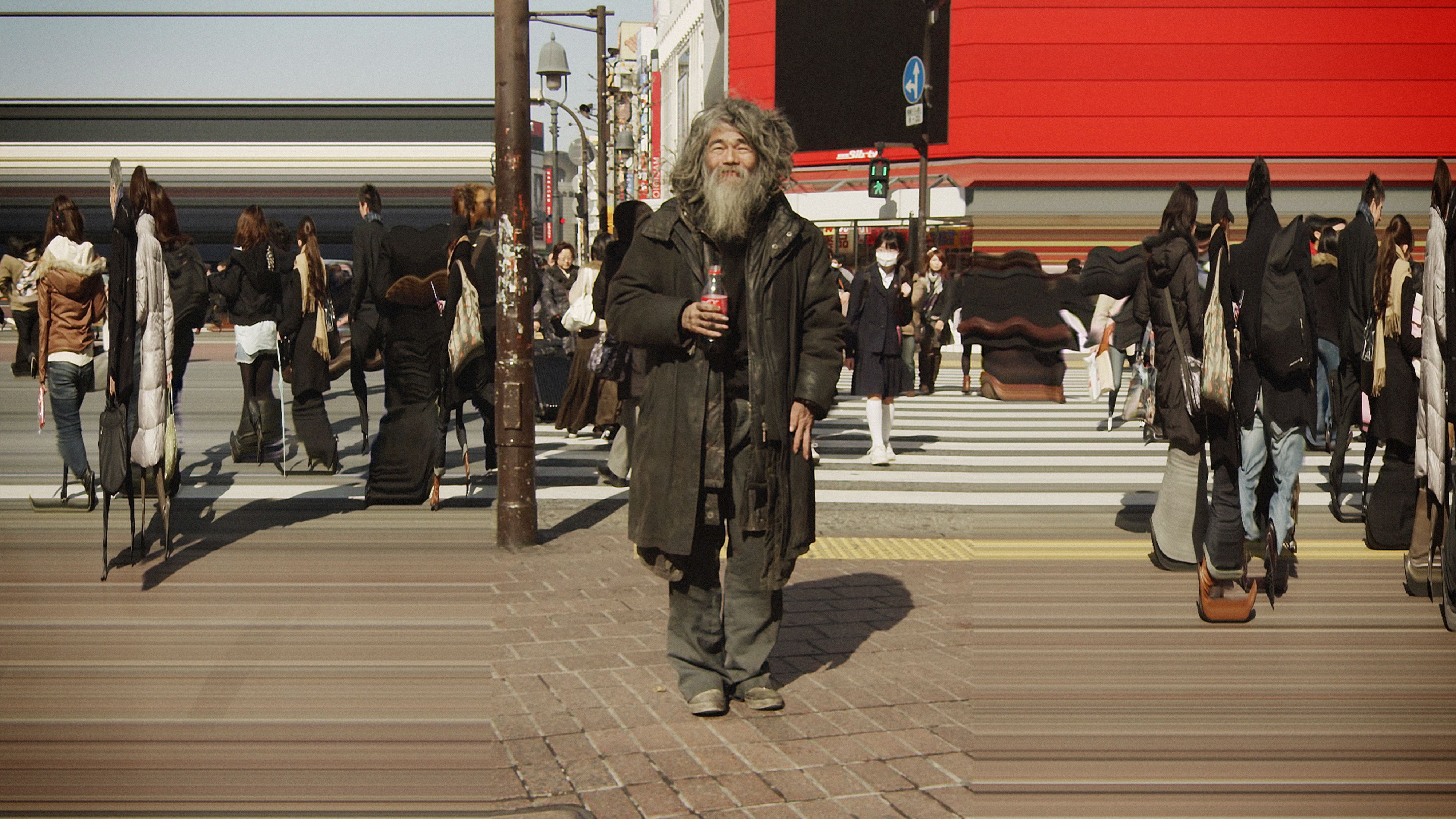 A still image of a man in a black puffer coat with grey hair and a long beard, standing in the middle of a crossing, with distorted images on either side of the frame of people walking.
