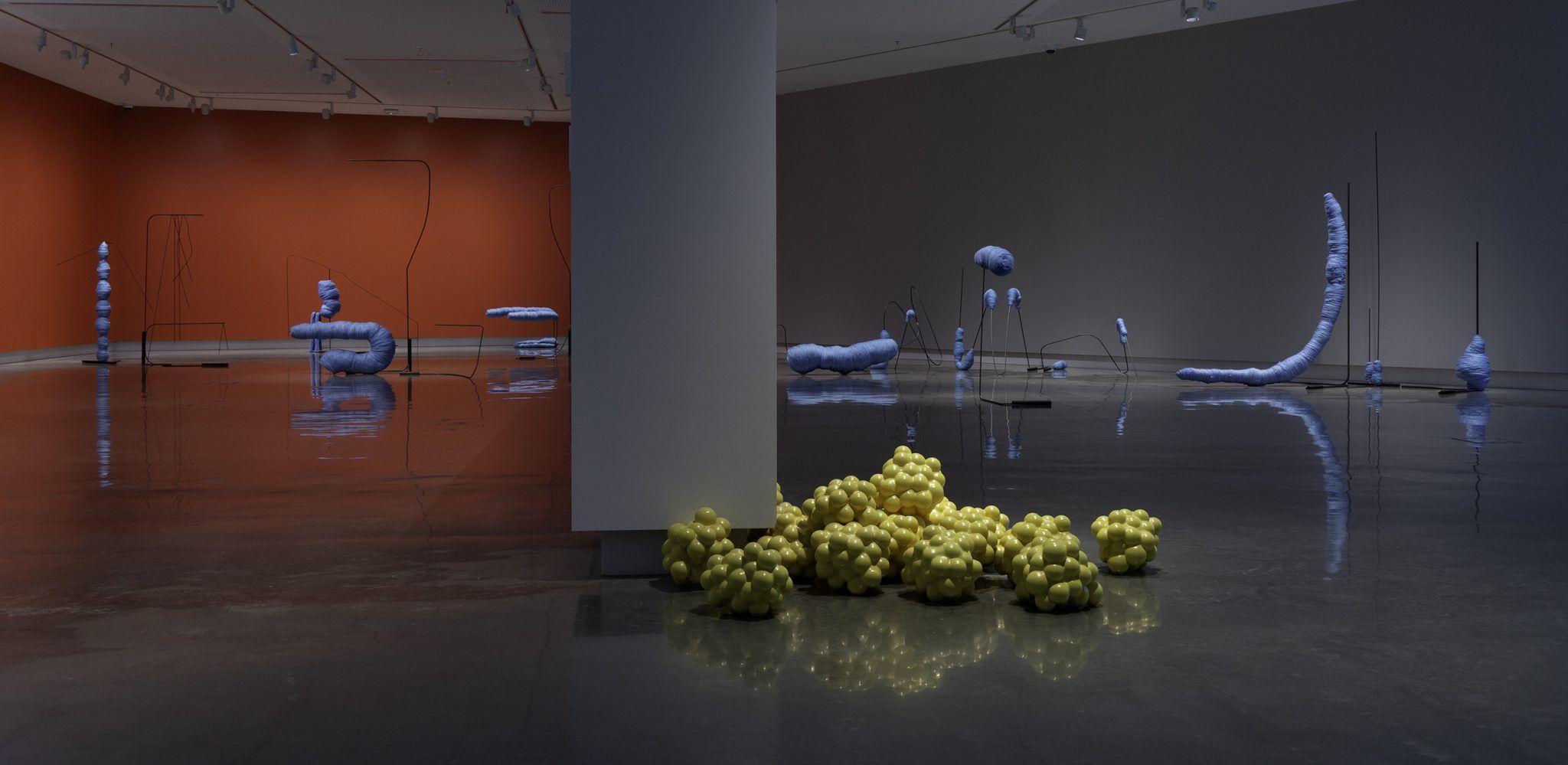 A contemporary art gallery with scattered abstract sculptures: pale purple organic shapes on thin stands and a cluster of yellow, grape-like forms on the floor near a central pillar. The walls are orange and gray, and the floor is polished concrete.